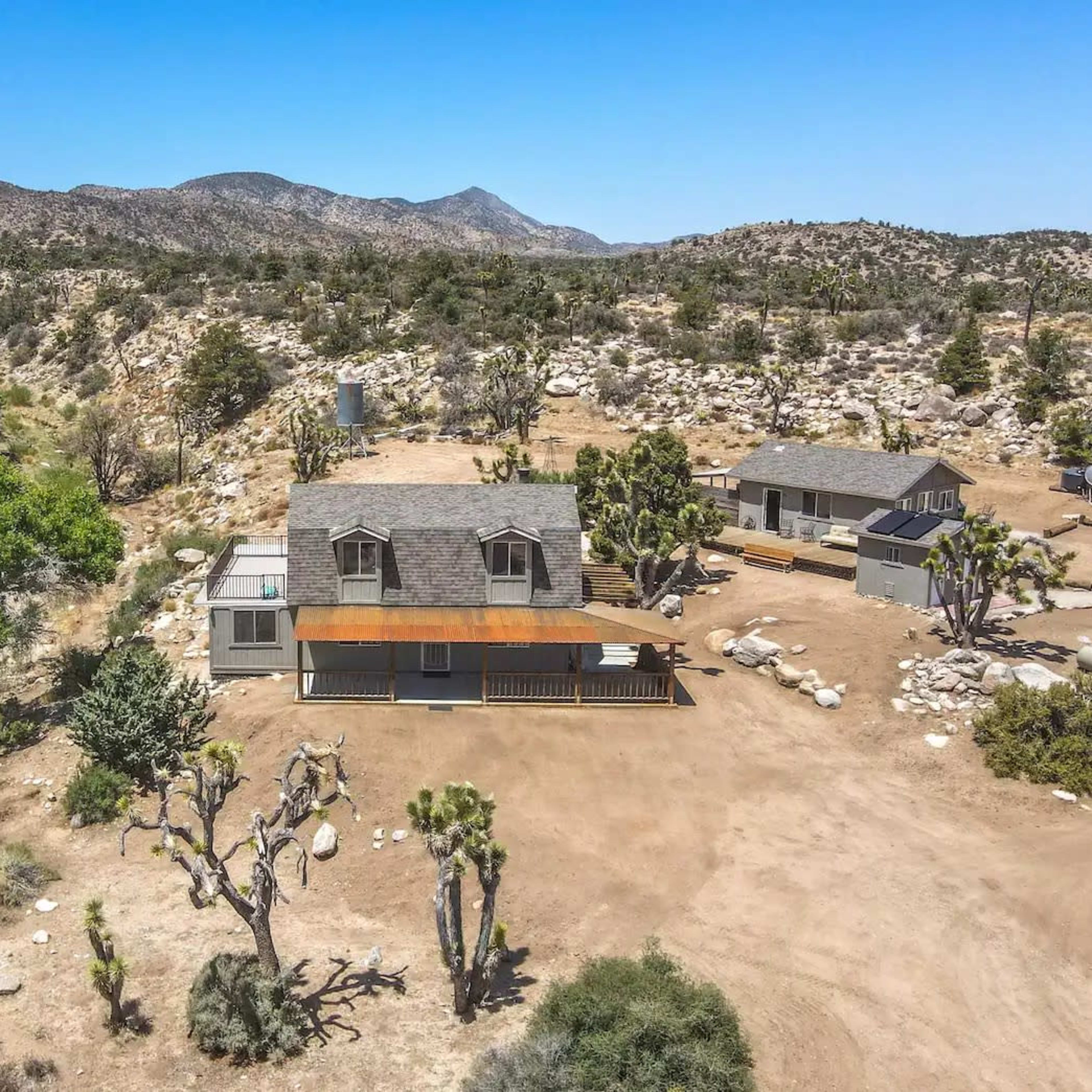 A single-story house with an extended deck surrounded by sparse vegetation and rocky terrain in a mountainous area.