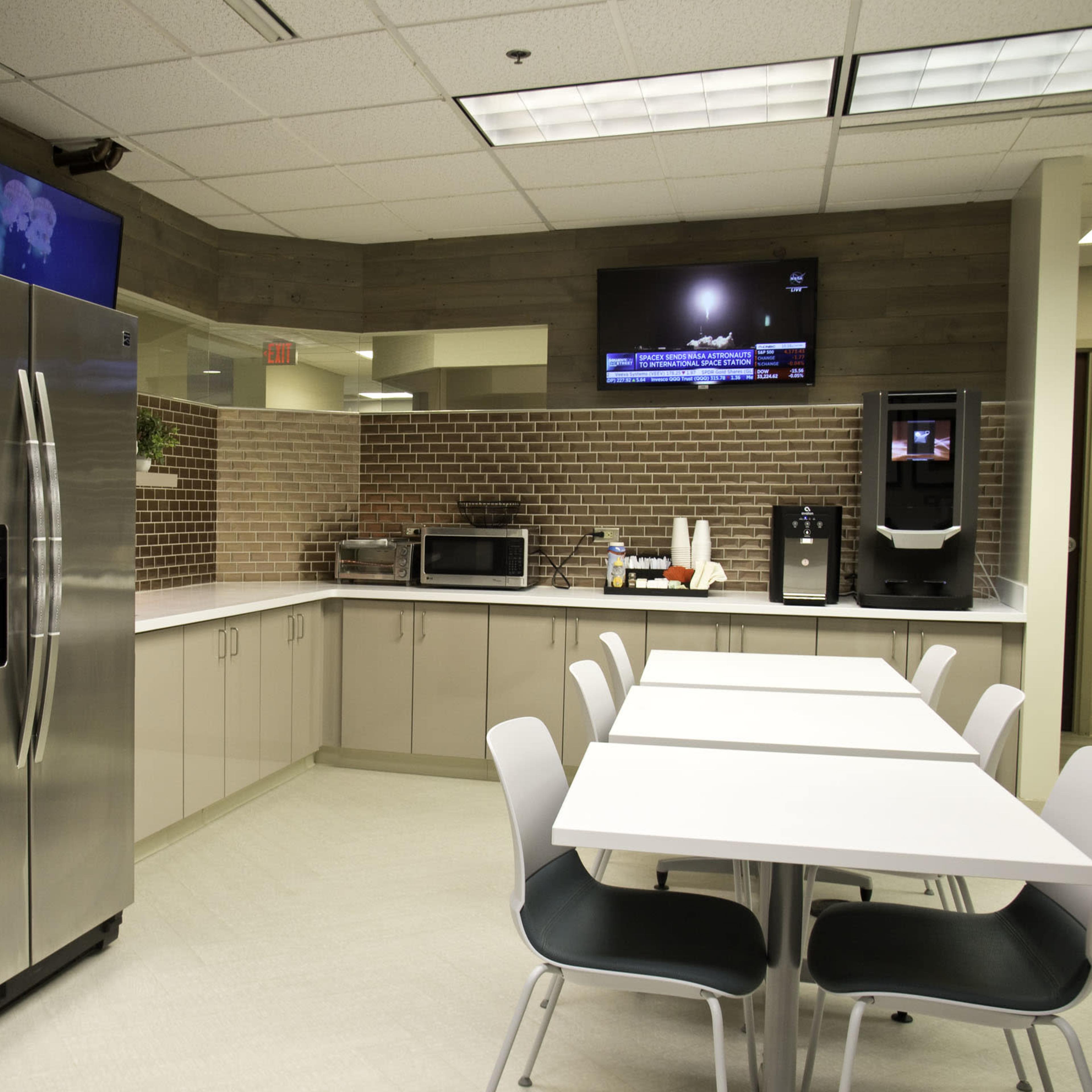 A modern break room featuring a vending machine, refrigerator, microwave, and a seating area with white tables and black chairs.