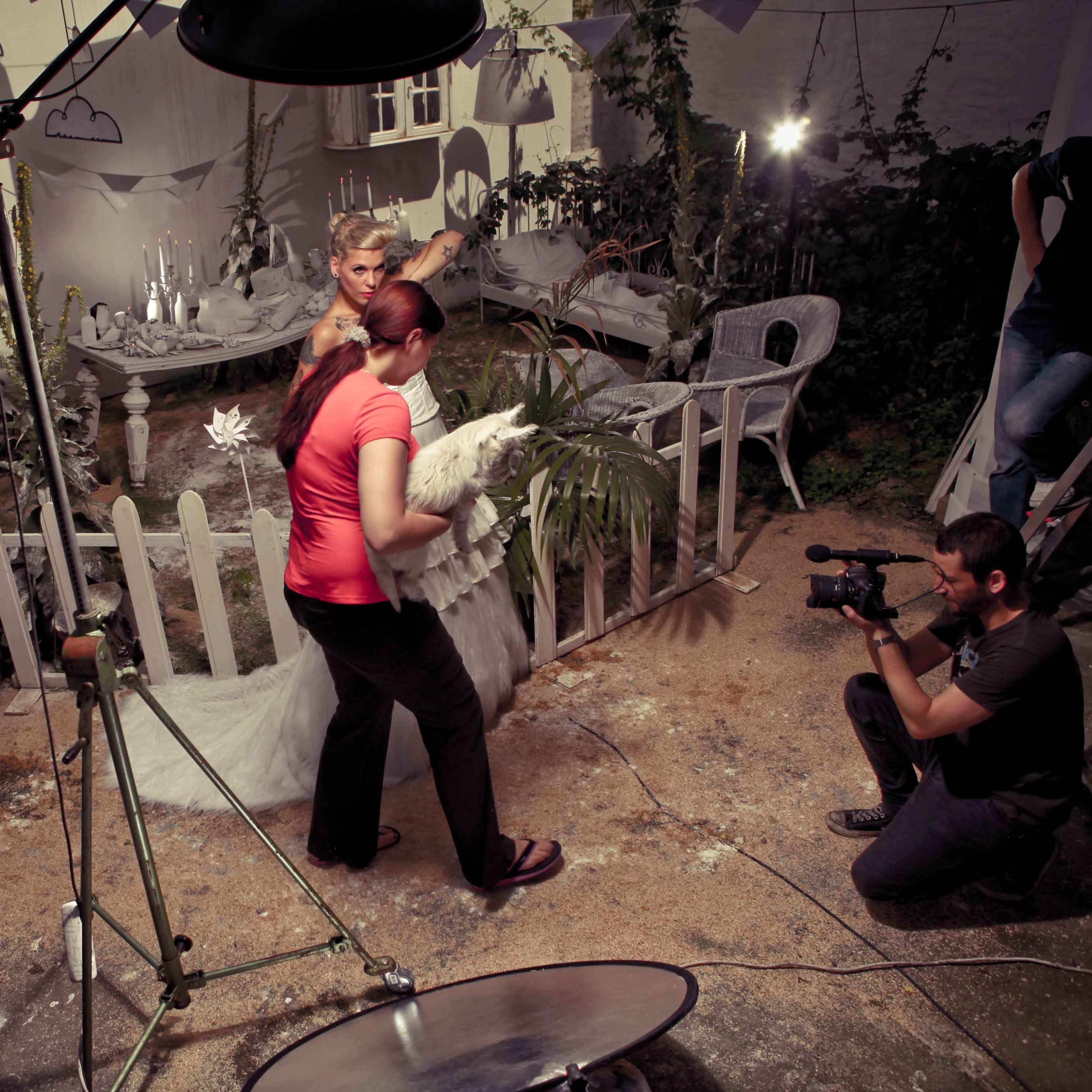 A woman carries a chicken while a group of people and lighting equipment set up for a photography shoot in a garden-like space.