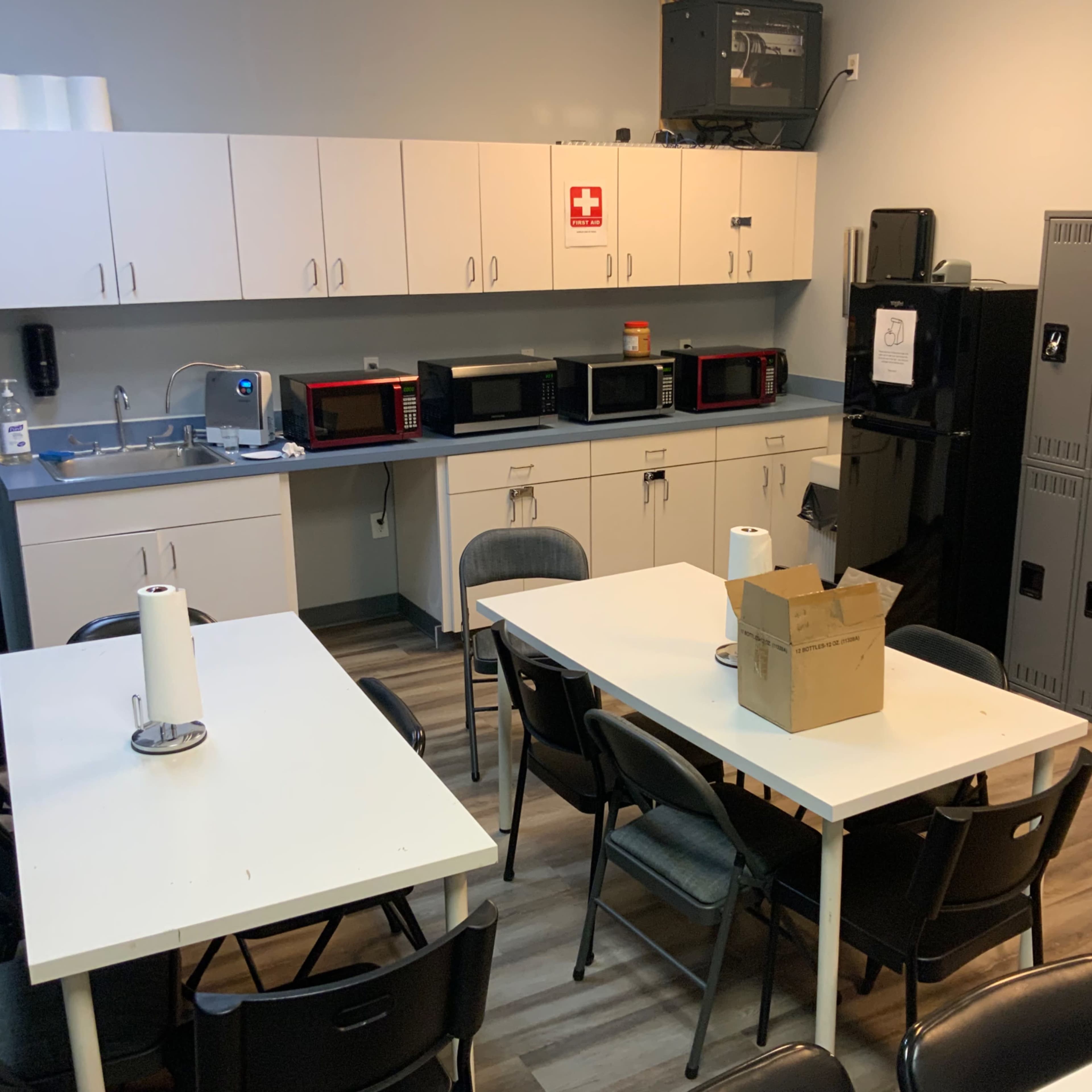 A break room with white tables and chairs, gray lockers, a kitchenette with multiple microwaves, and a refrigerator.