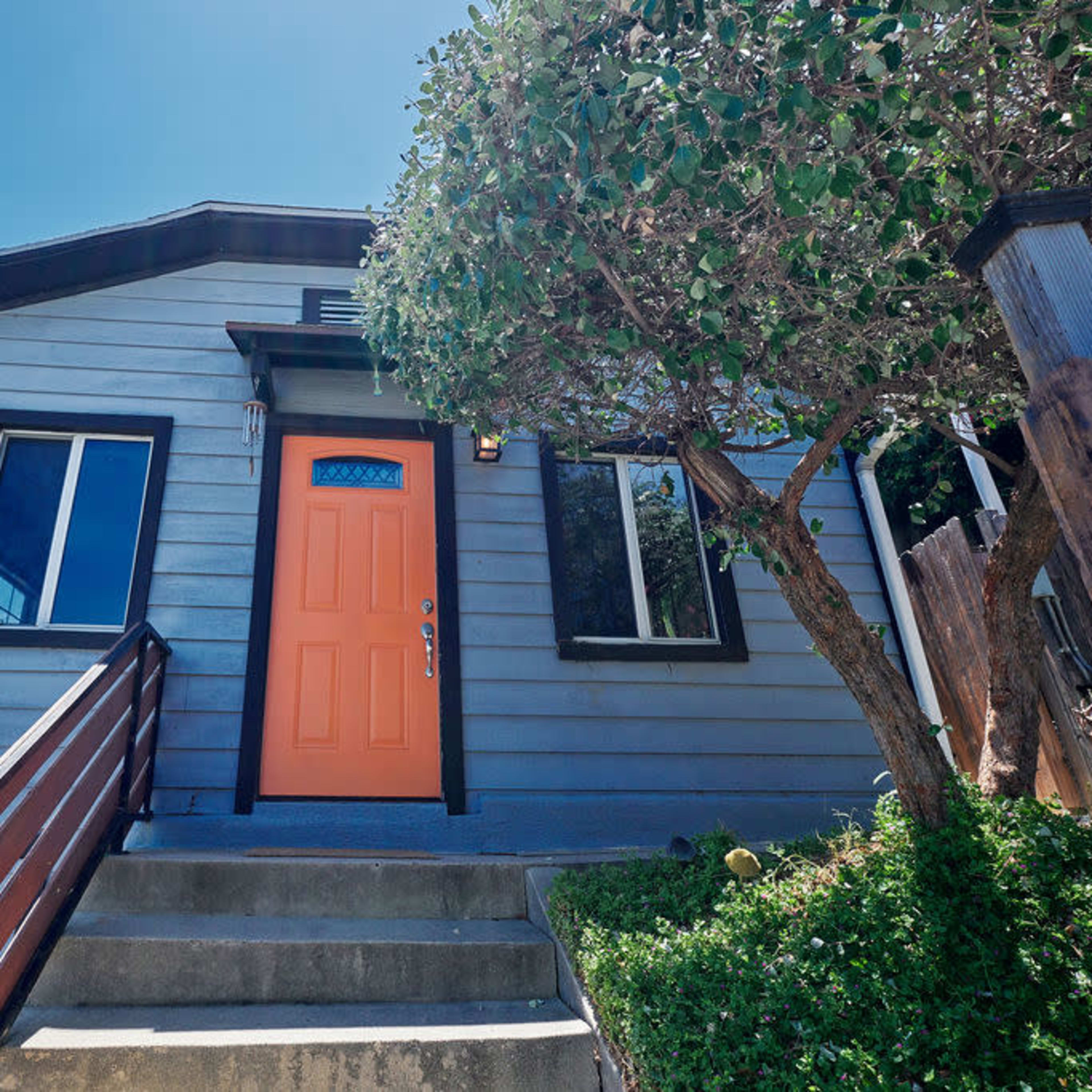 A light blue house features an orange front door and a sloped entrance with wooden steps.