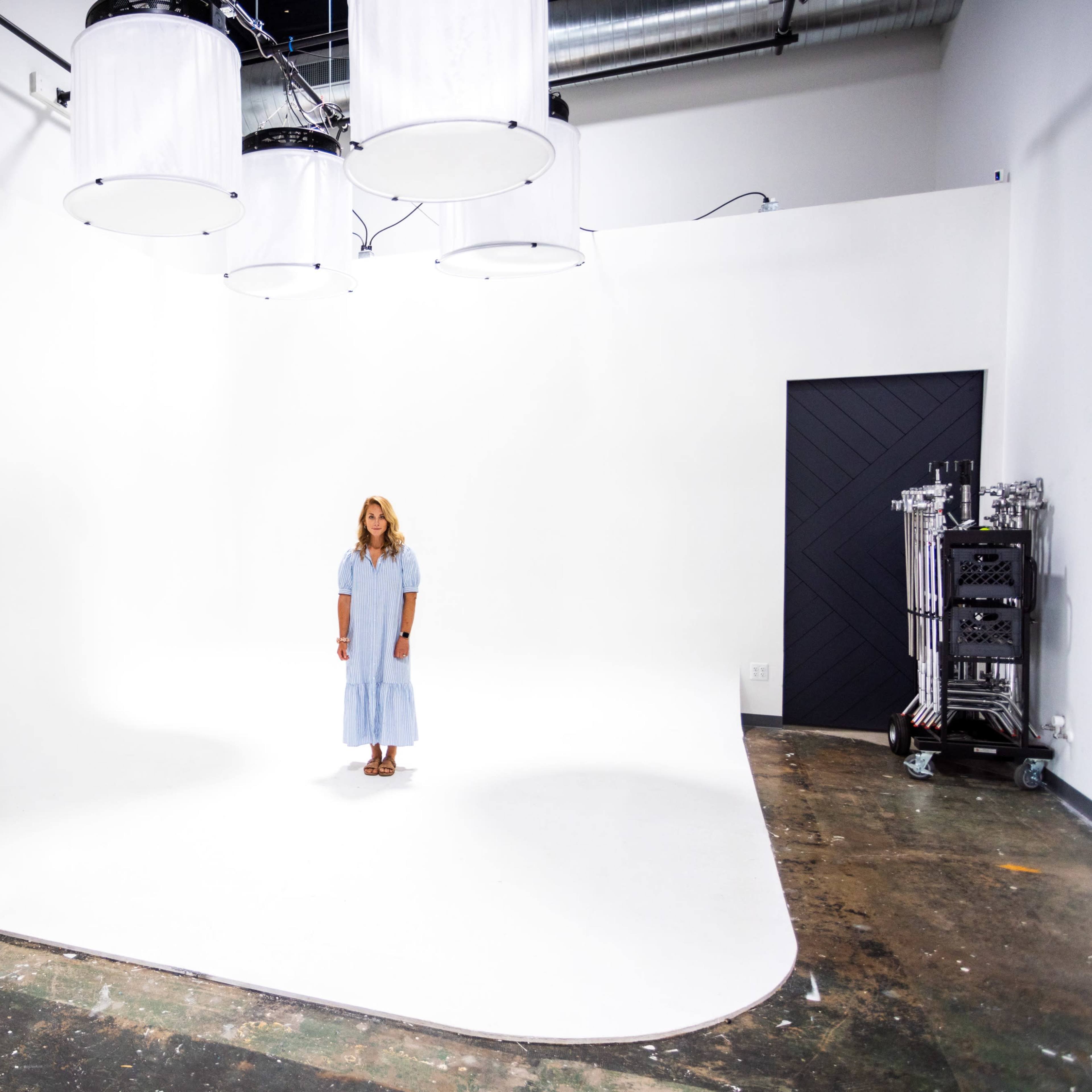 A woman stands alone on a white backdrop inside a spacious studio with overhead lights and a dark door.