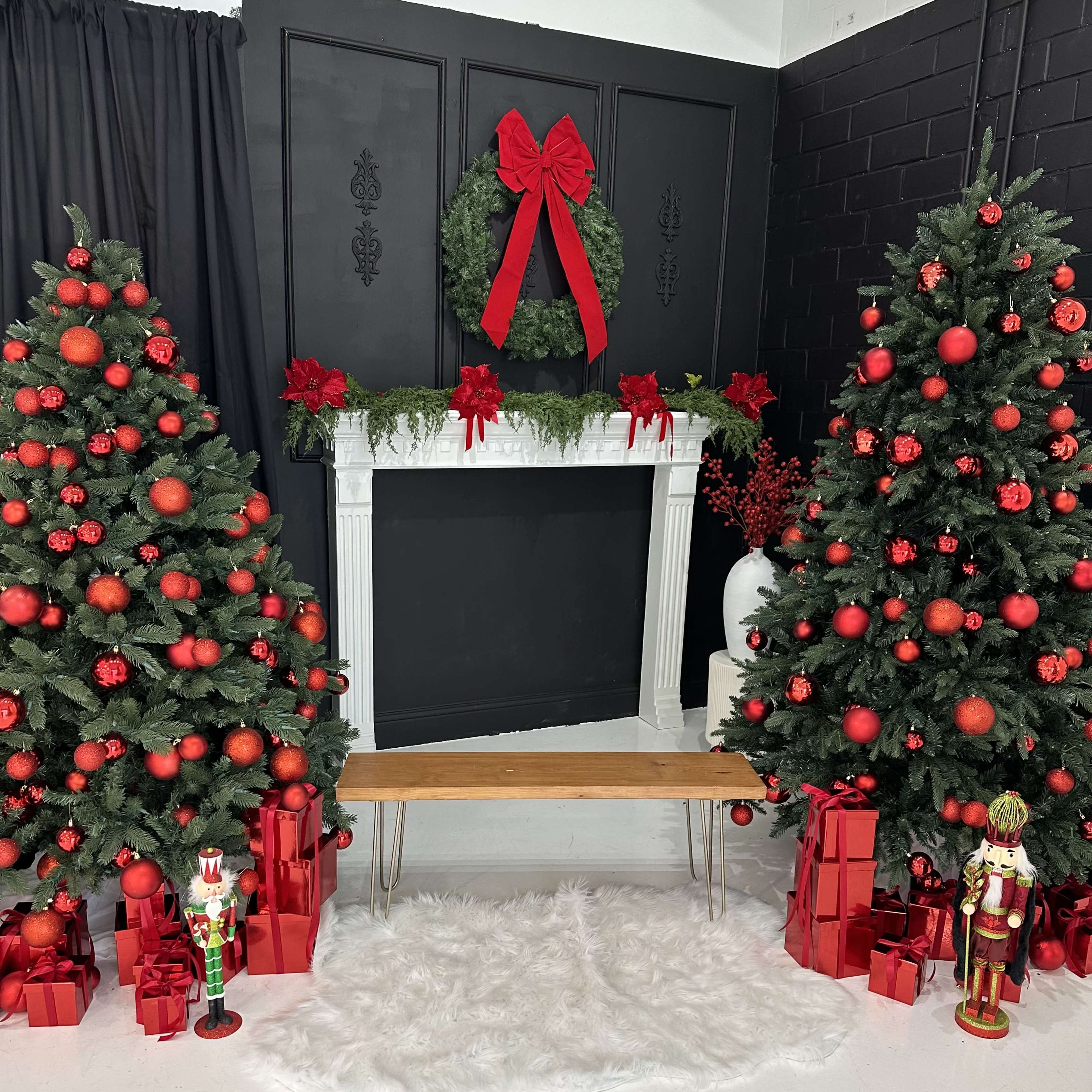 A decorated holiday scene with two Christmas trees adorned with red ornaments flanking a white faux fur rug and a wooden bench in front of a black wall with a white mantelpiece draped in greenery.