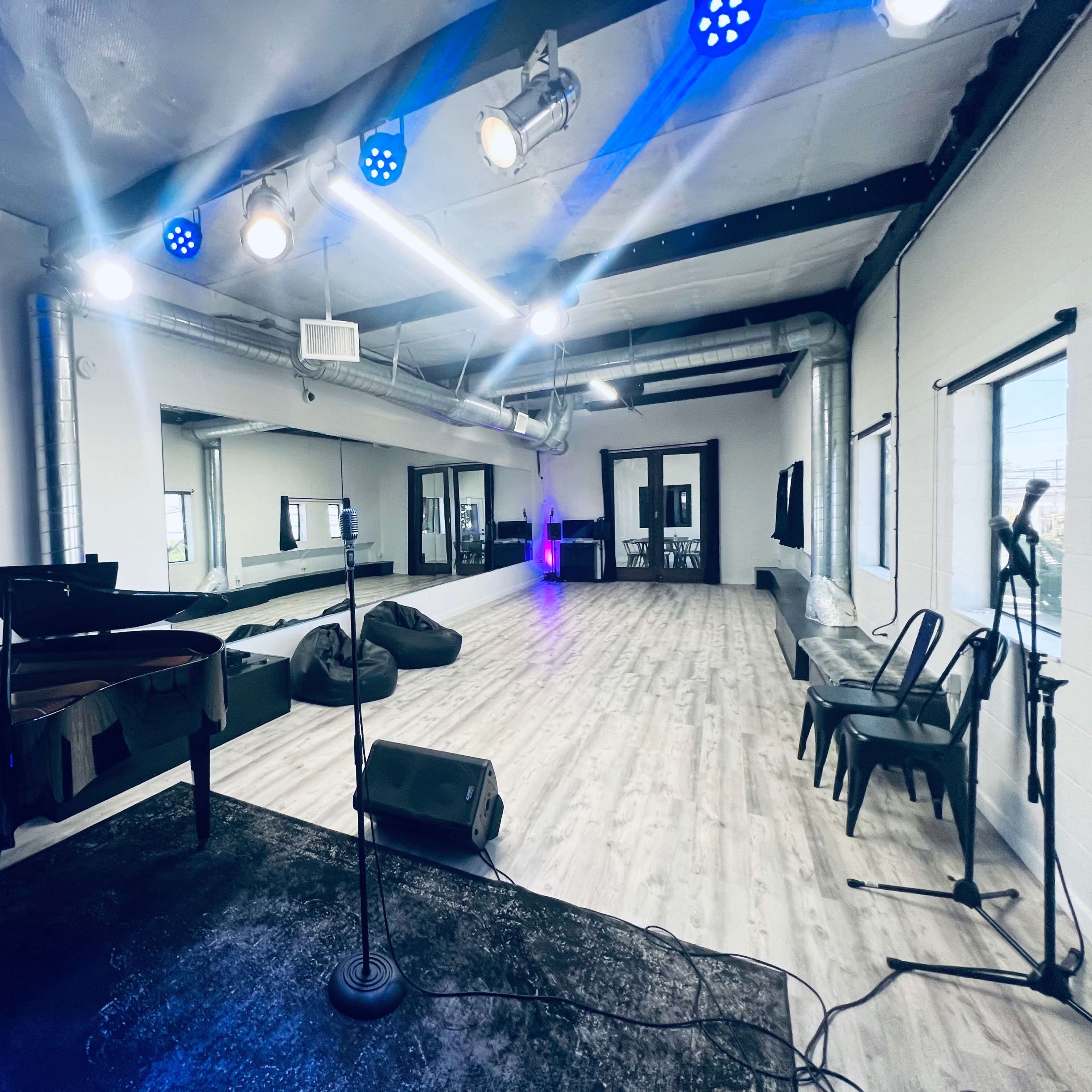 A spacious performance studio with a grand piano, a microphone stand, and several chairs arranged on a wooden floor, illuminated by stage lights.
