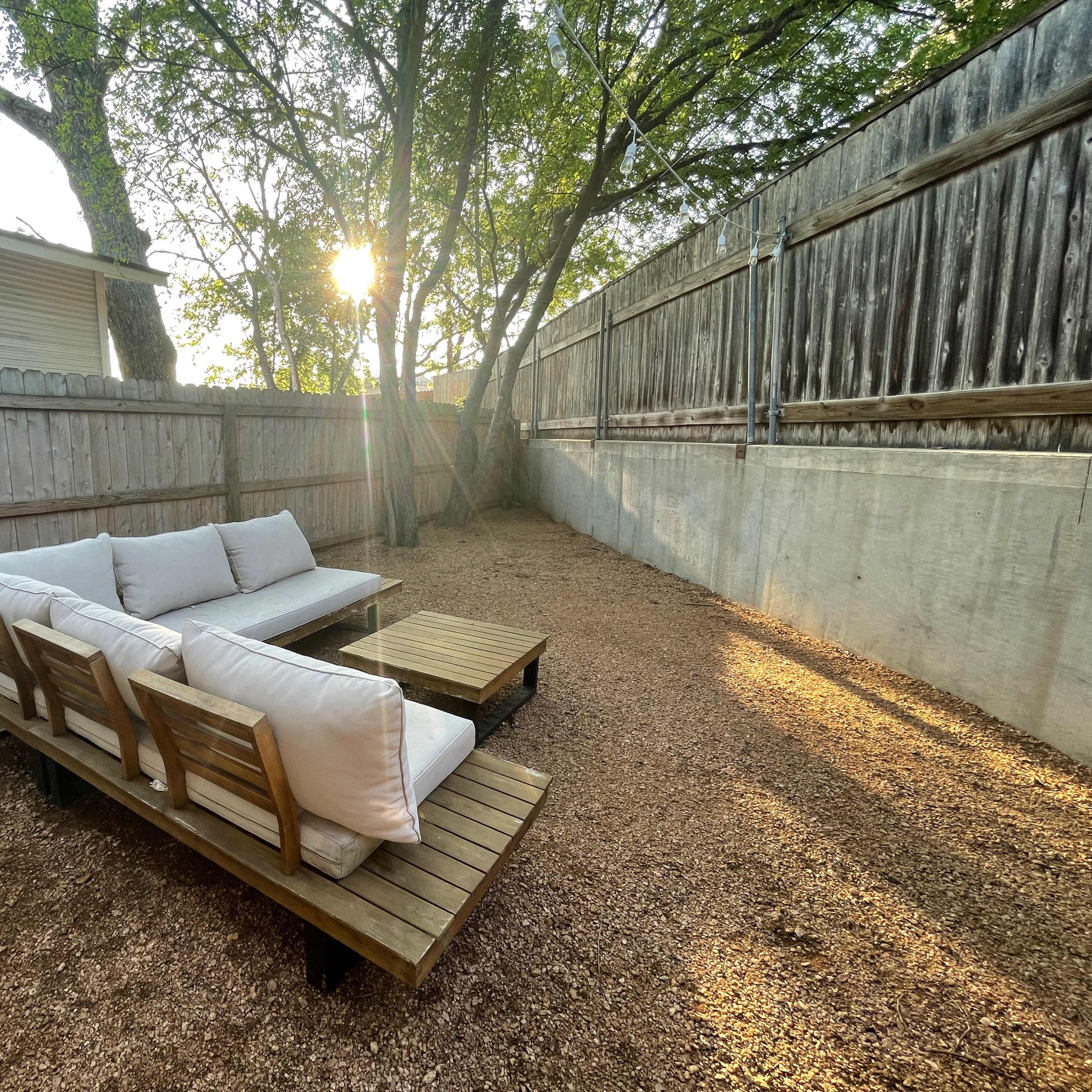 A wooden seating area with white cushions is set on gravel in a backyard, illuminated by sunlight filtering through the trees.