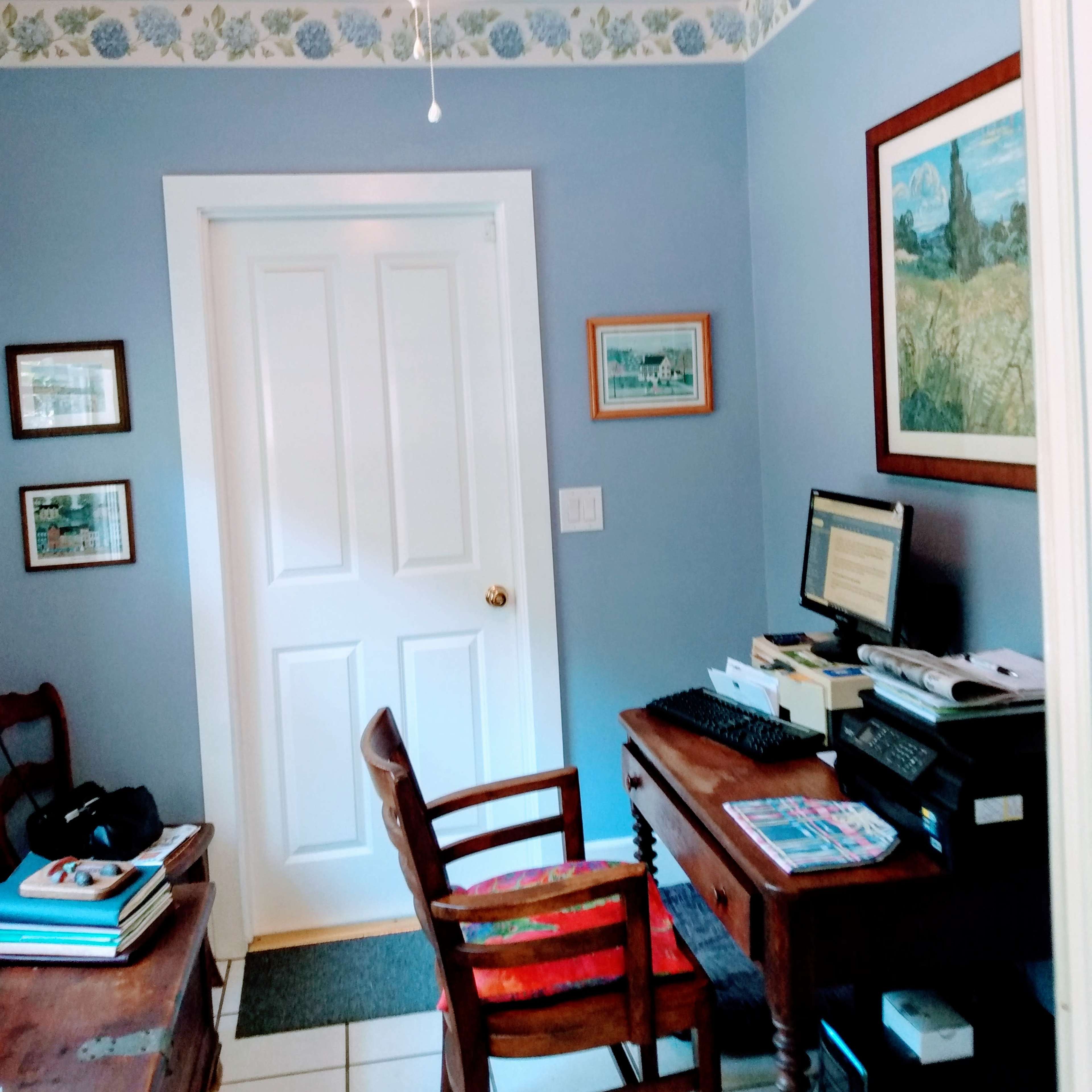 A home office with a wooden desk and chair, a computer, a decorated wall with art, and a door leading to another room.