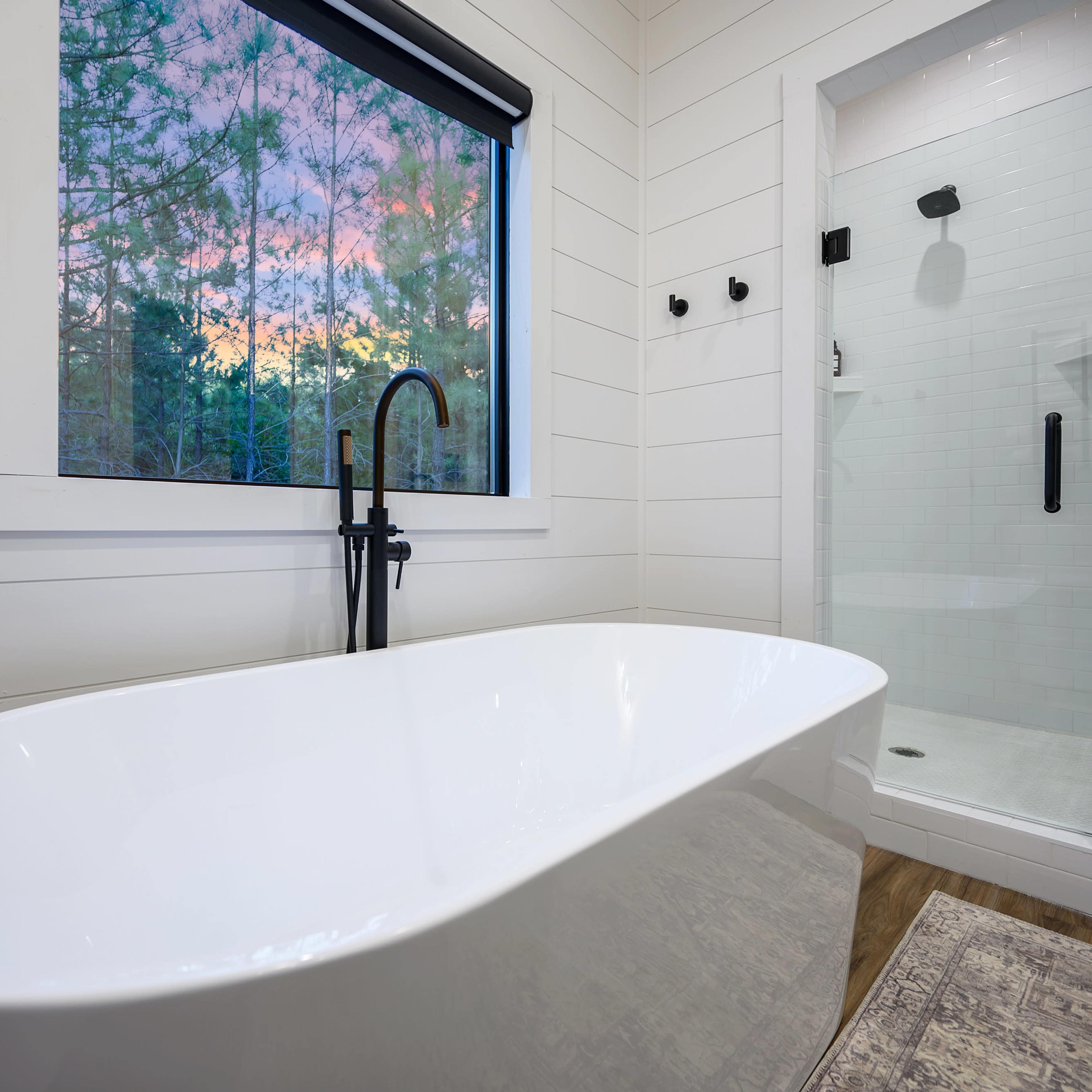 A modern bathroom features a freestanding white bathtub next to a glass shower enclosure, with a large window revealing a colorful sunset.