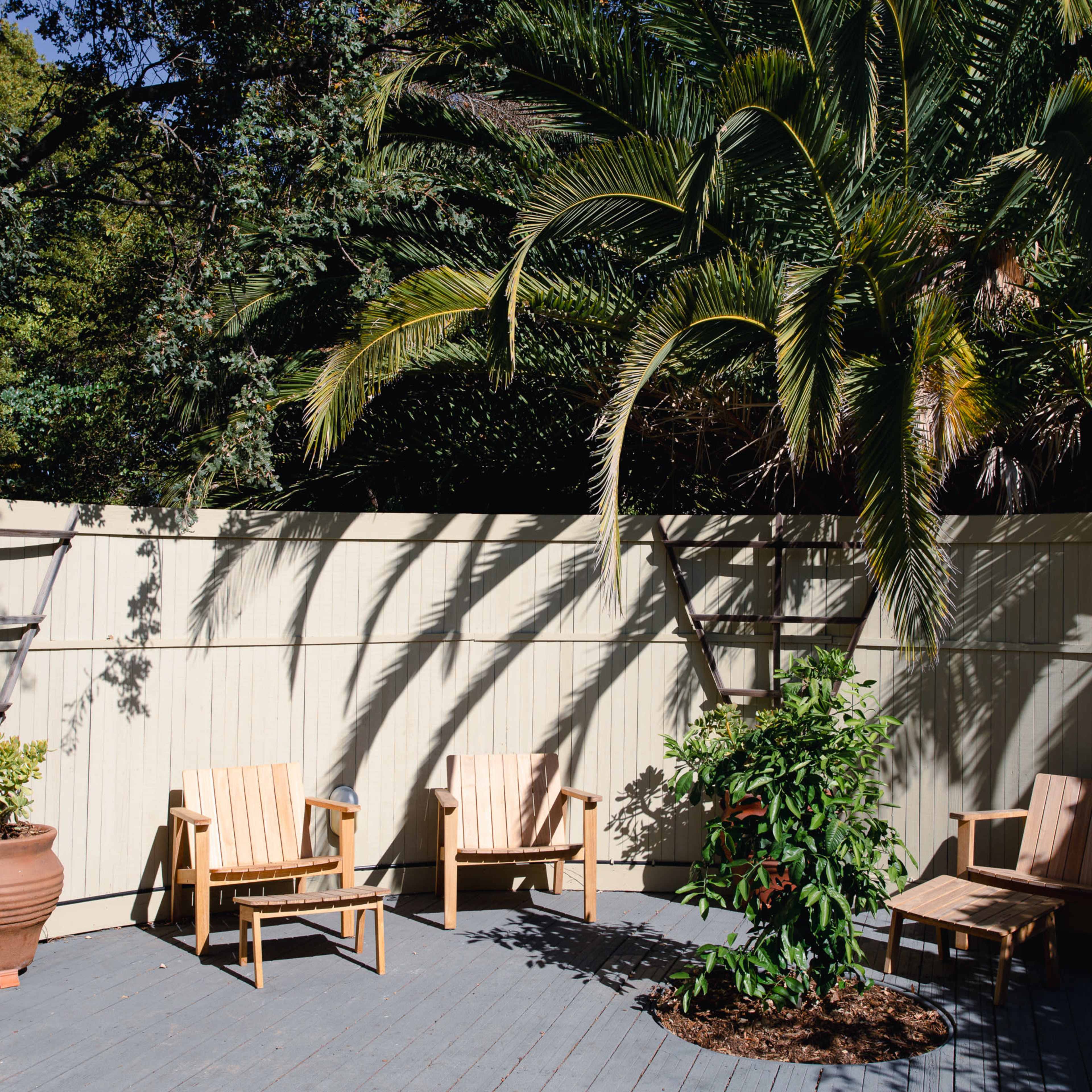 A small outdoor seating area features wooden chairs arranged around a potted plant, set against a pale fence and shaded by palm trees.