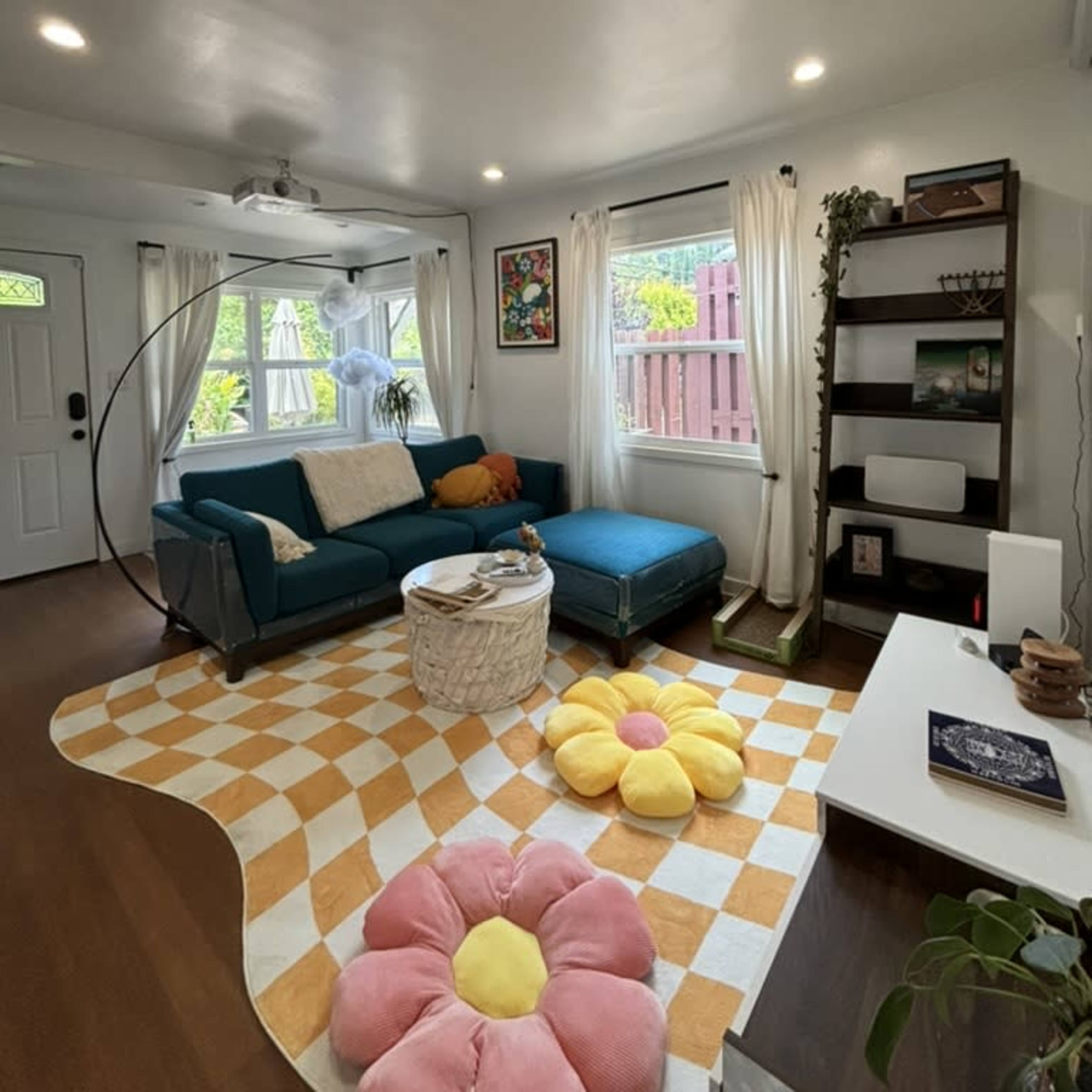 A cozy living room featuring a teal sectional sofa, a round coffee table, and a large flower-shaped cushion on a checkered area rug.