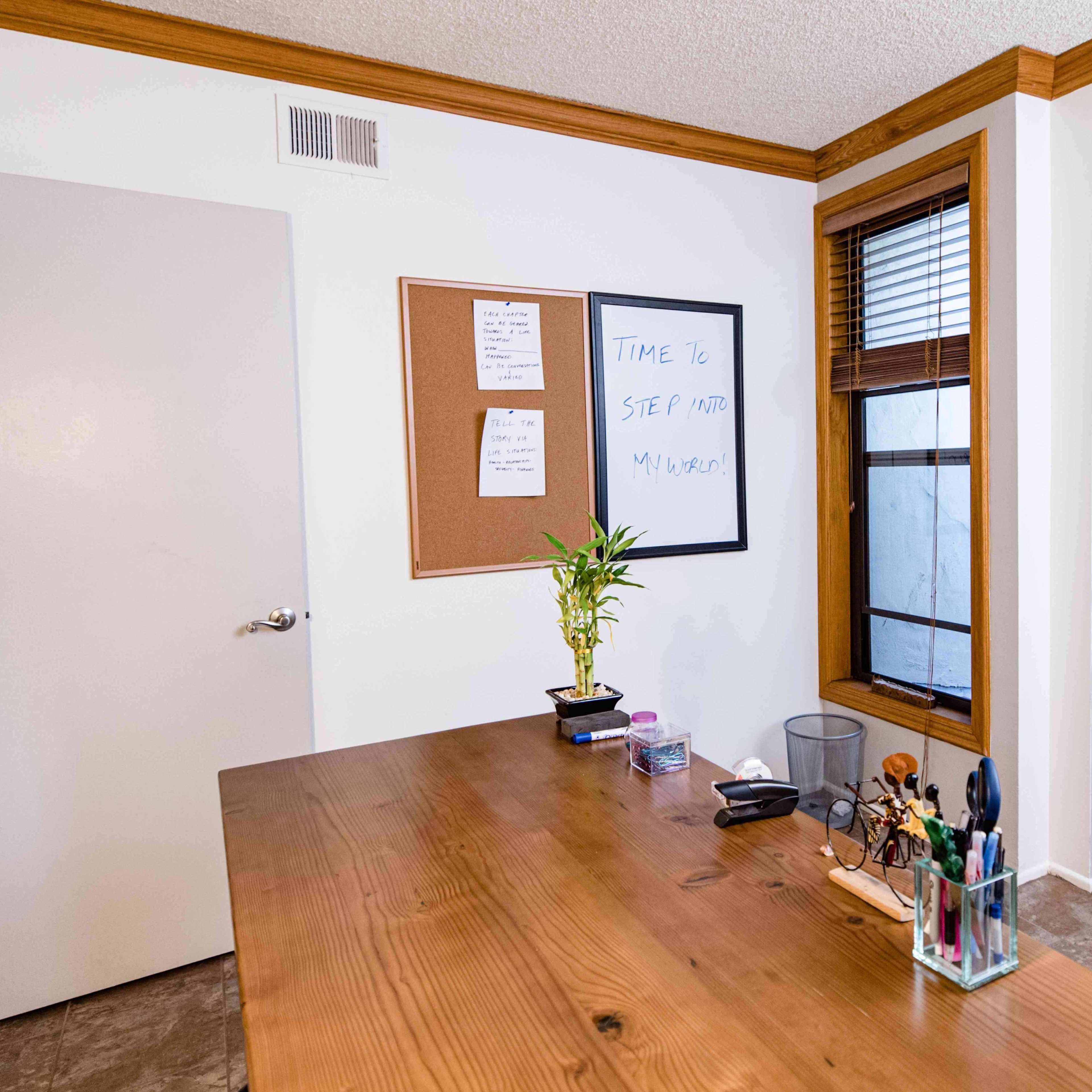 A wooden desk is positioned facing a corkboard and a whiteboard on a wall, with a closed door and a window nearby.