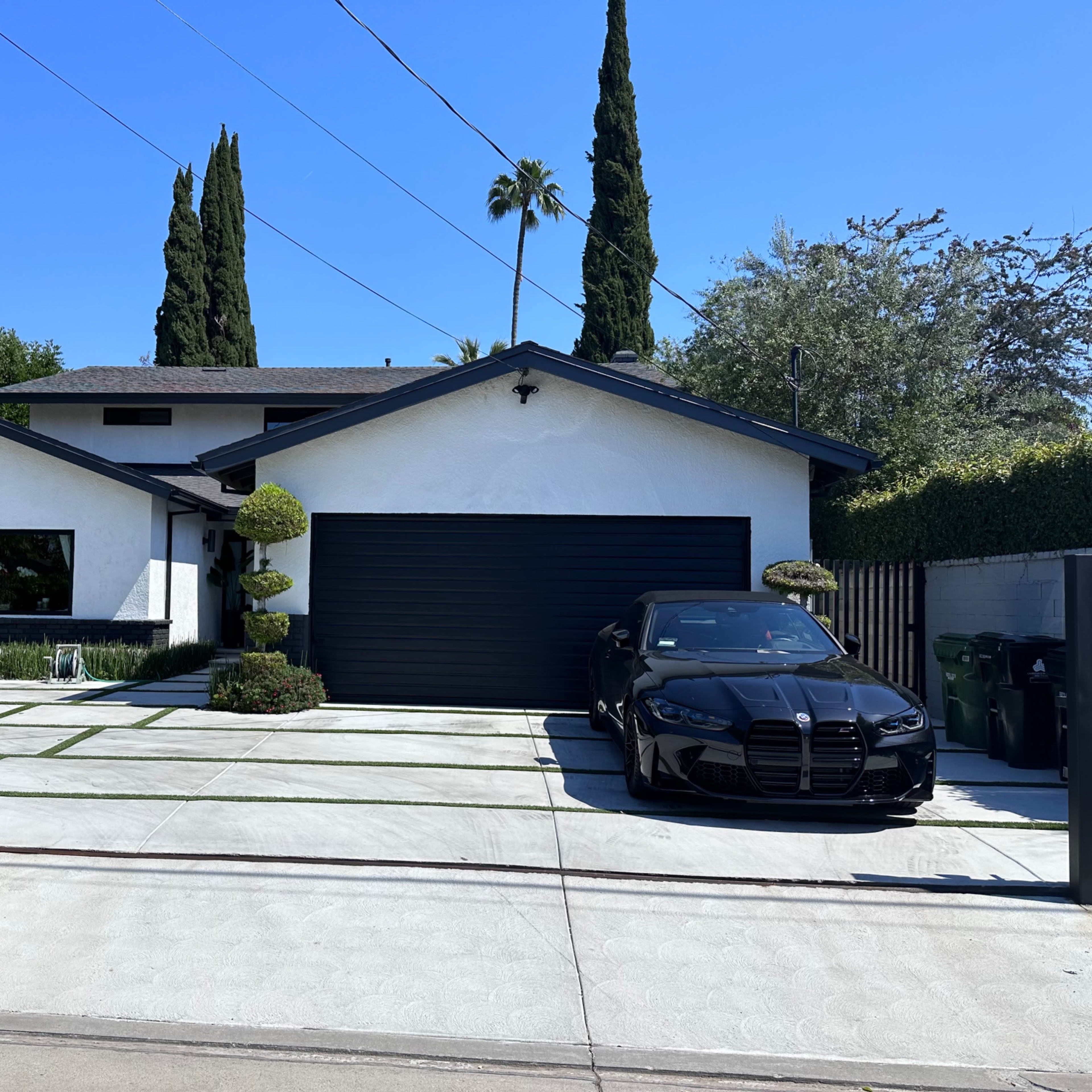 A modern white house with a black garage door and a black car parked in front, framed by neatly trimmed shrubs and palm trees.