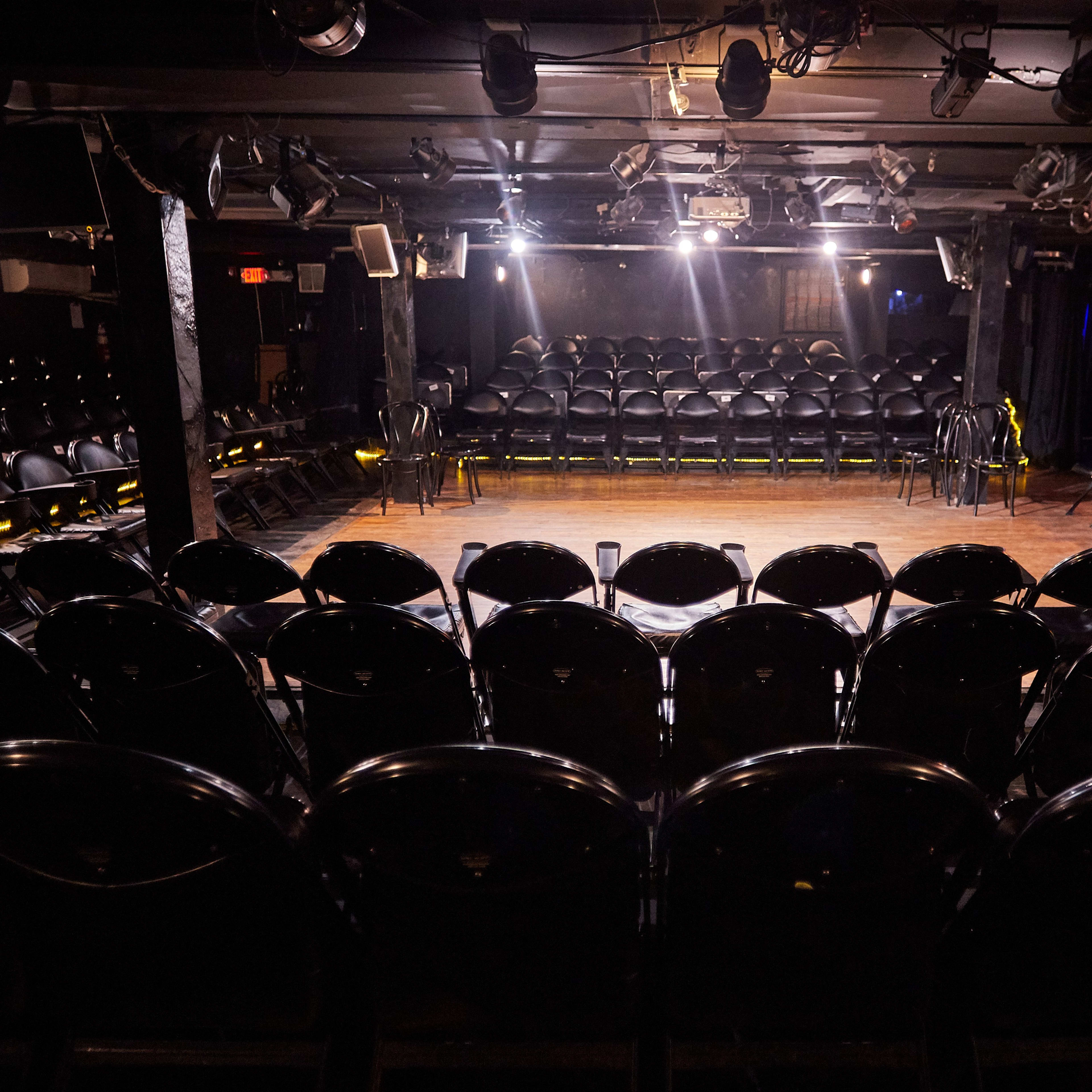 A dimly lit comedy club features a wooden stage surrounded by rows of black chairs.