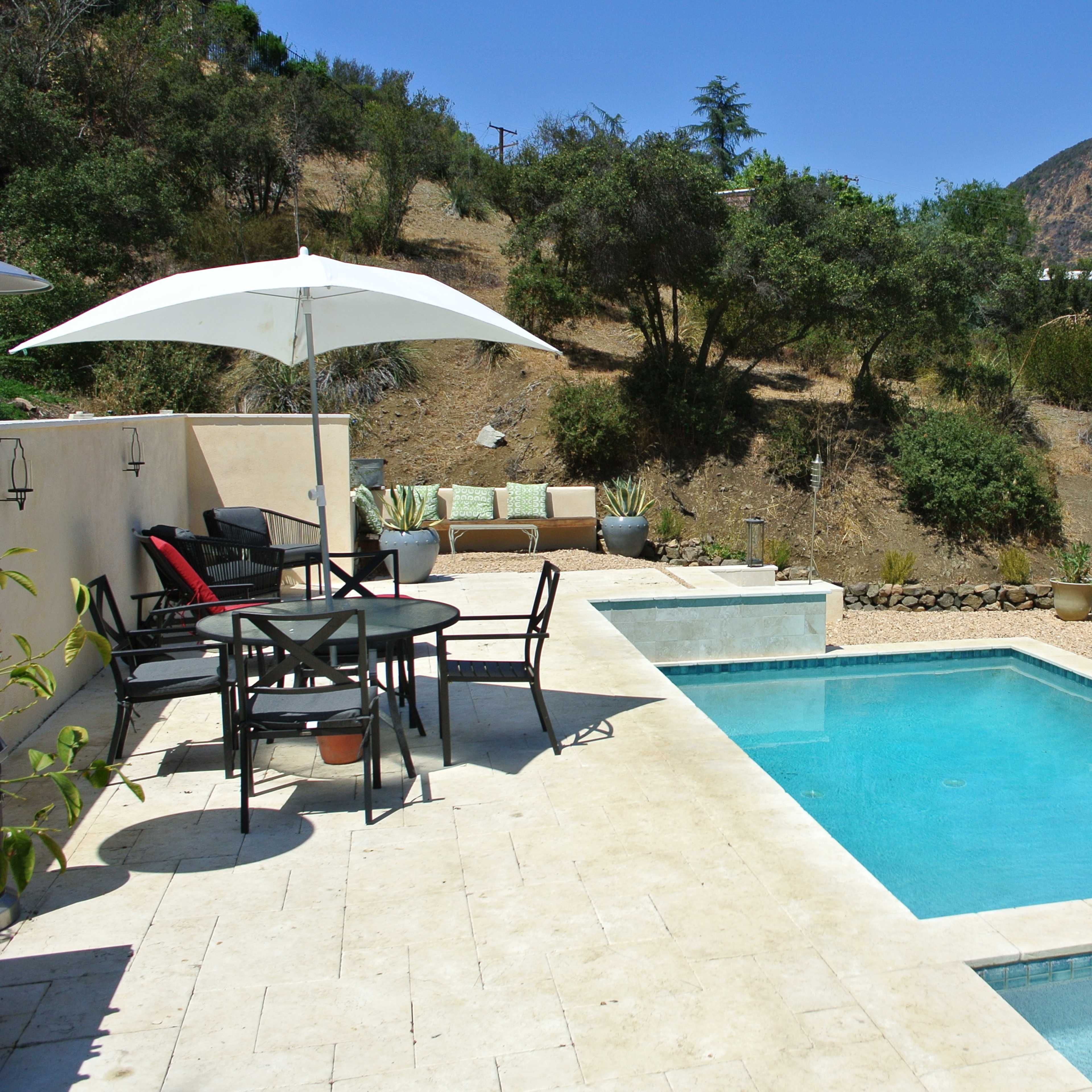 A modern backyard pool area with lounge chairs and patio furniture under white umbrellas, set against a backdrop of a hill.
