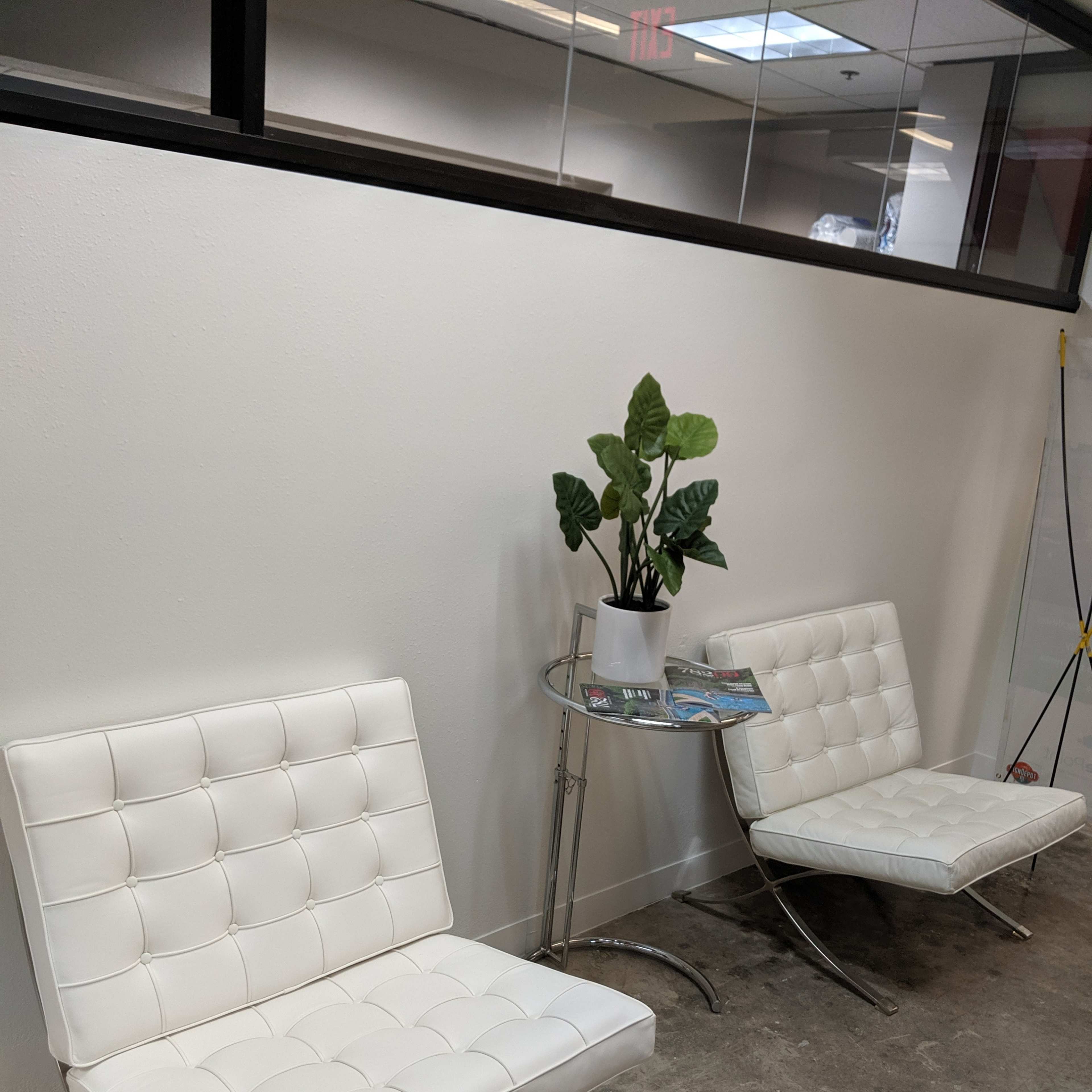 Two white chairs with tufted seats placed beside a small round table featuring a potted plant and a magazine, against a white wall in an office setting.