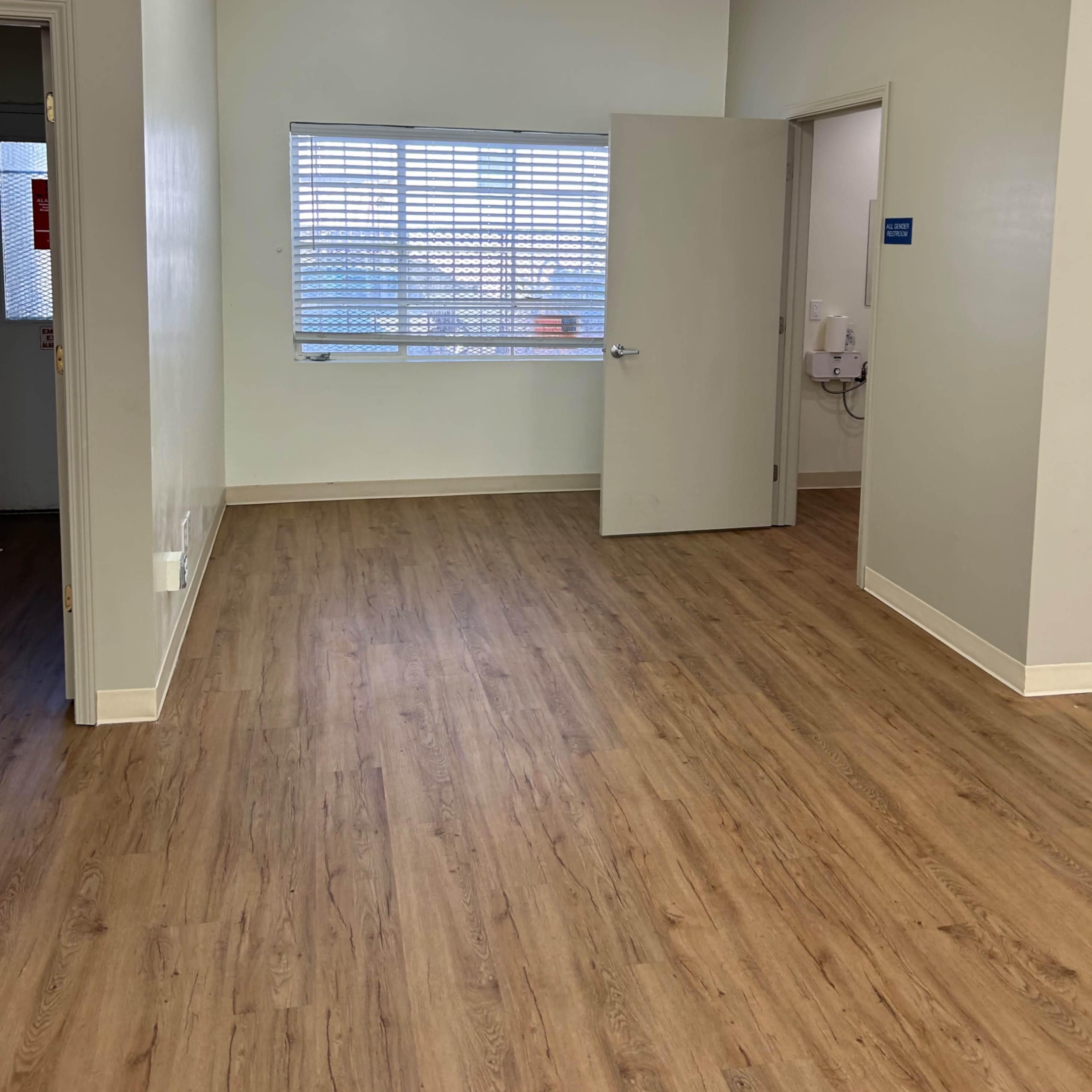 A well-lit empty room with wood-like flooring, a window with blinds, and an open door leading to another room.