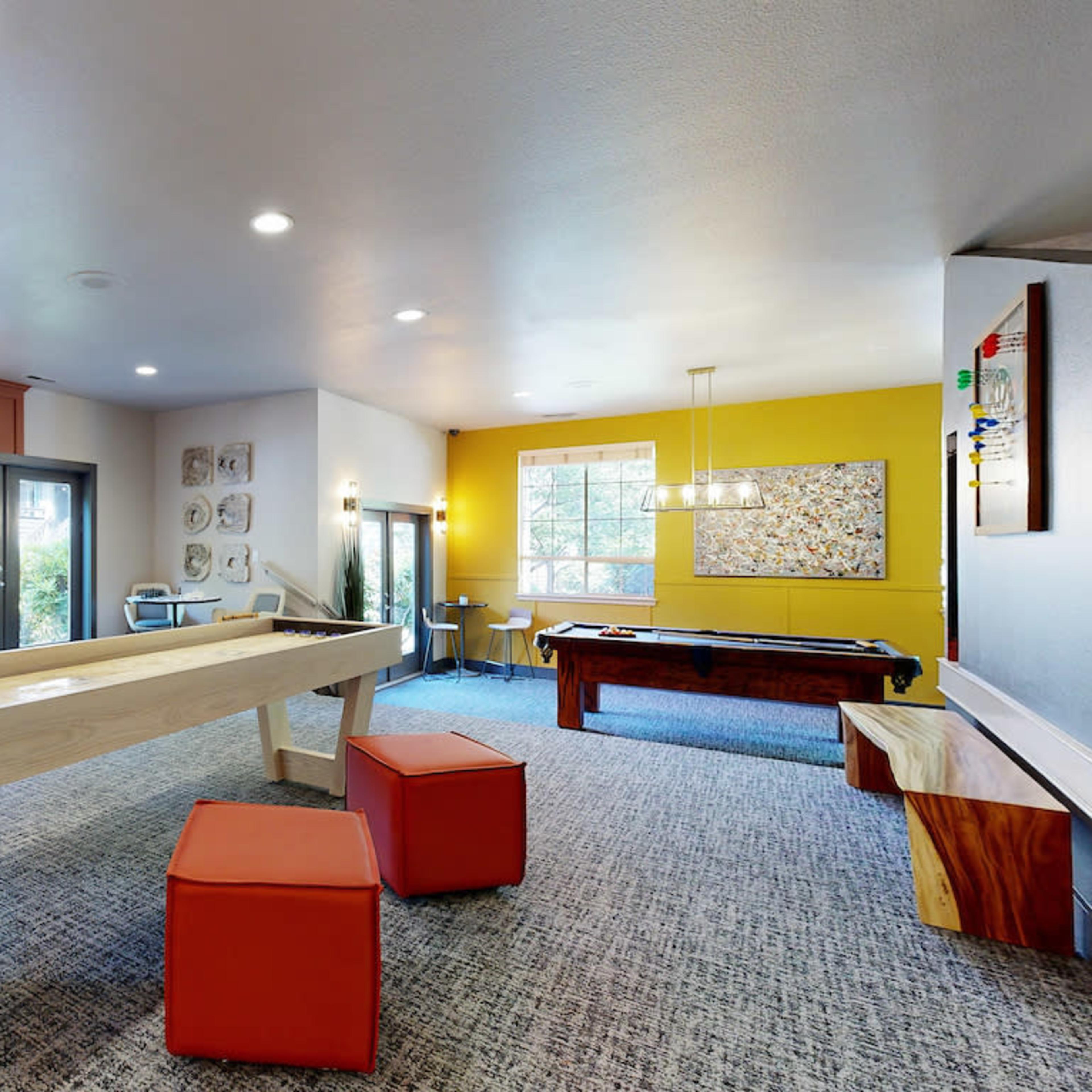 A modern recreational room featuring a fireplace, a large flat-screen TV, a shuffleboard table, and a billiards table, with colorful accents and seating areas.