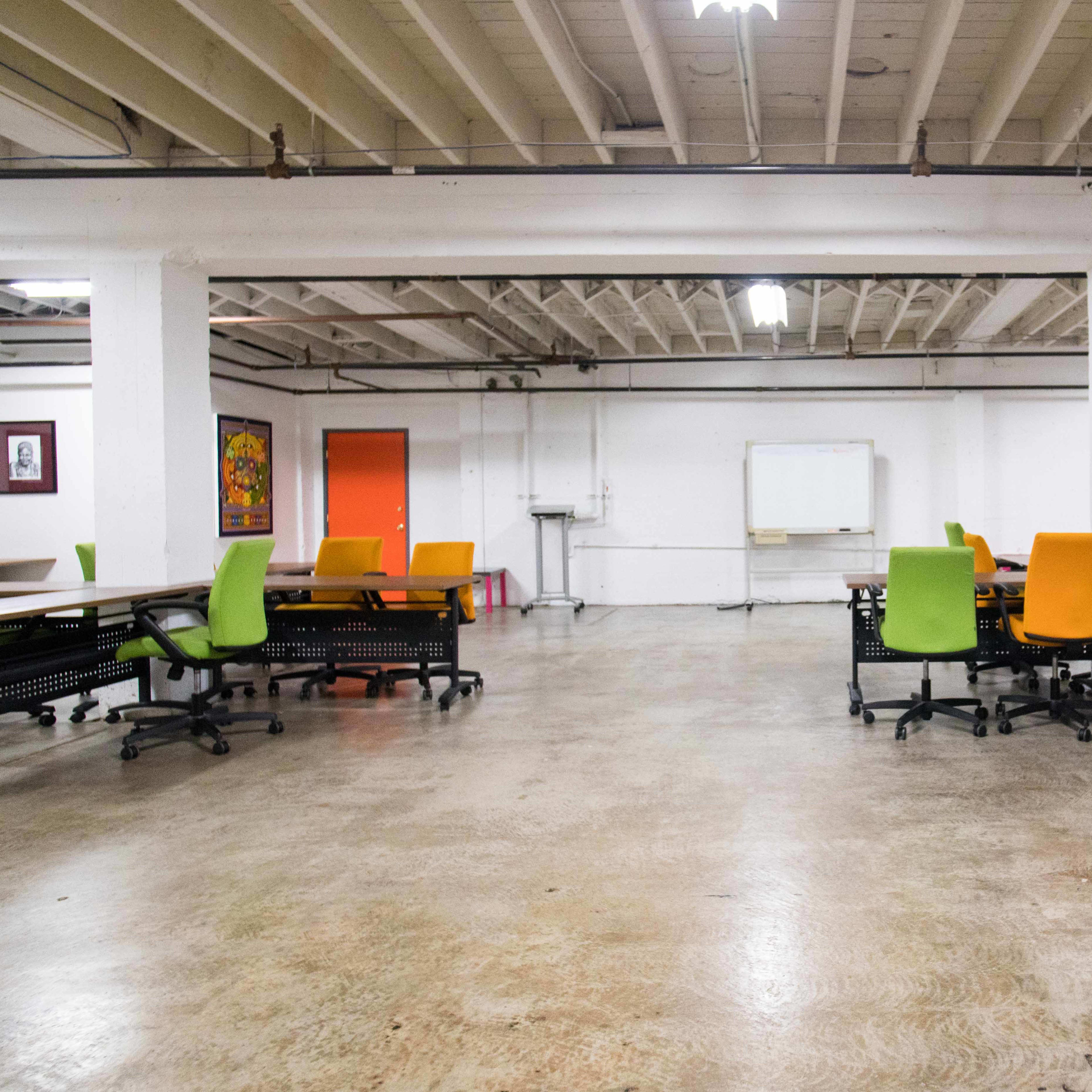 A spacious, empty office area featuring several desks with bright green and orange chairs, along with a whiteboard and orange door in the background.