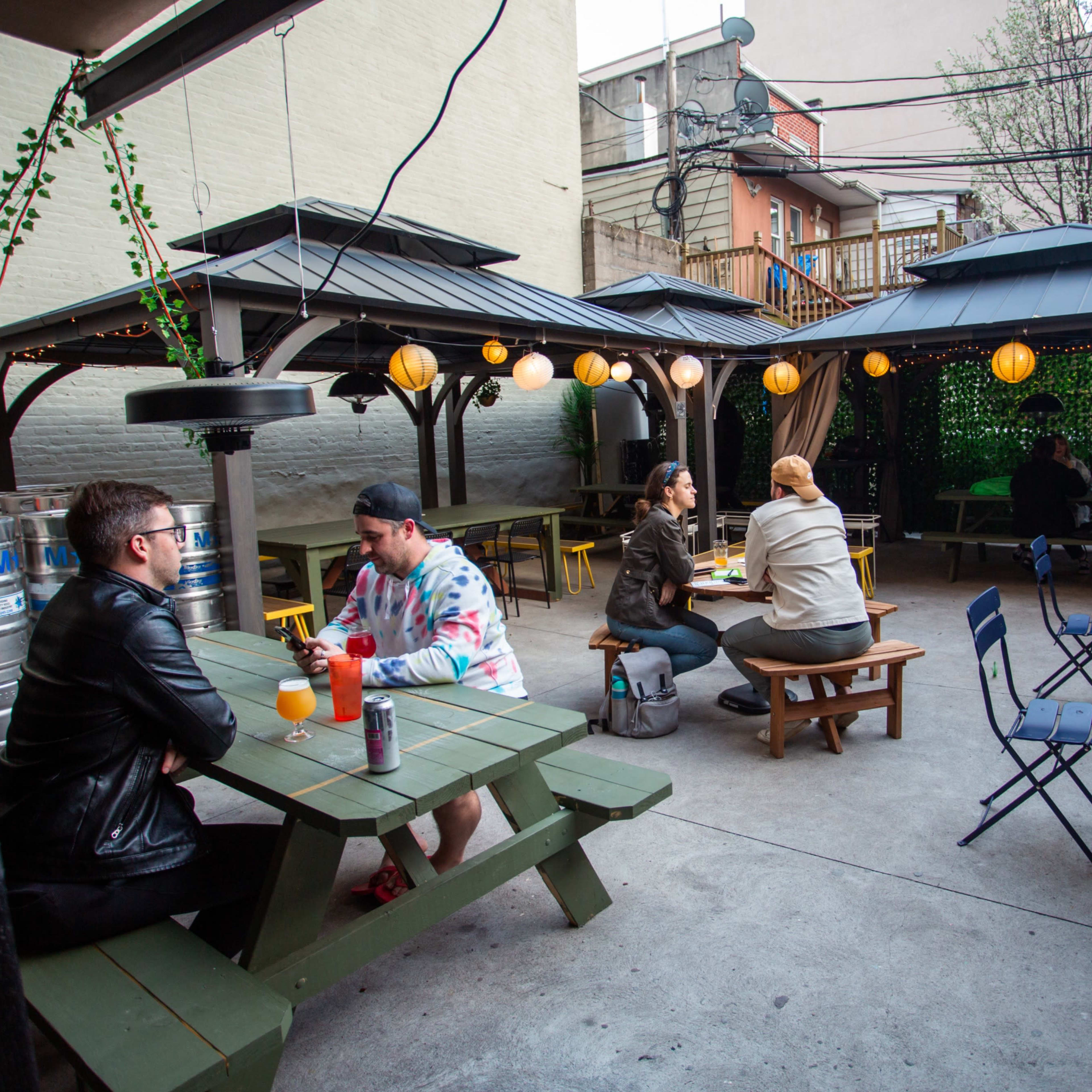 An outdoor patio area with several wooden picnic tables, string lights, and patrons sitting under covered gazebos.