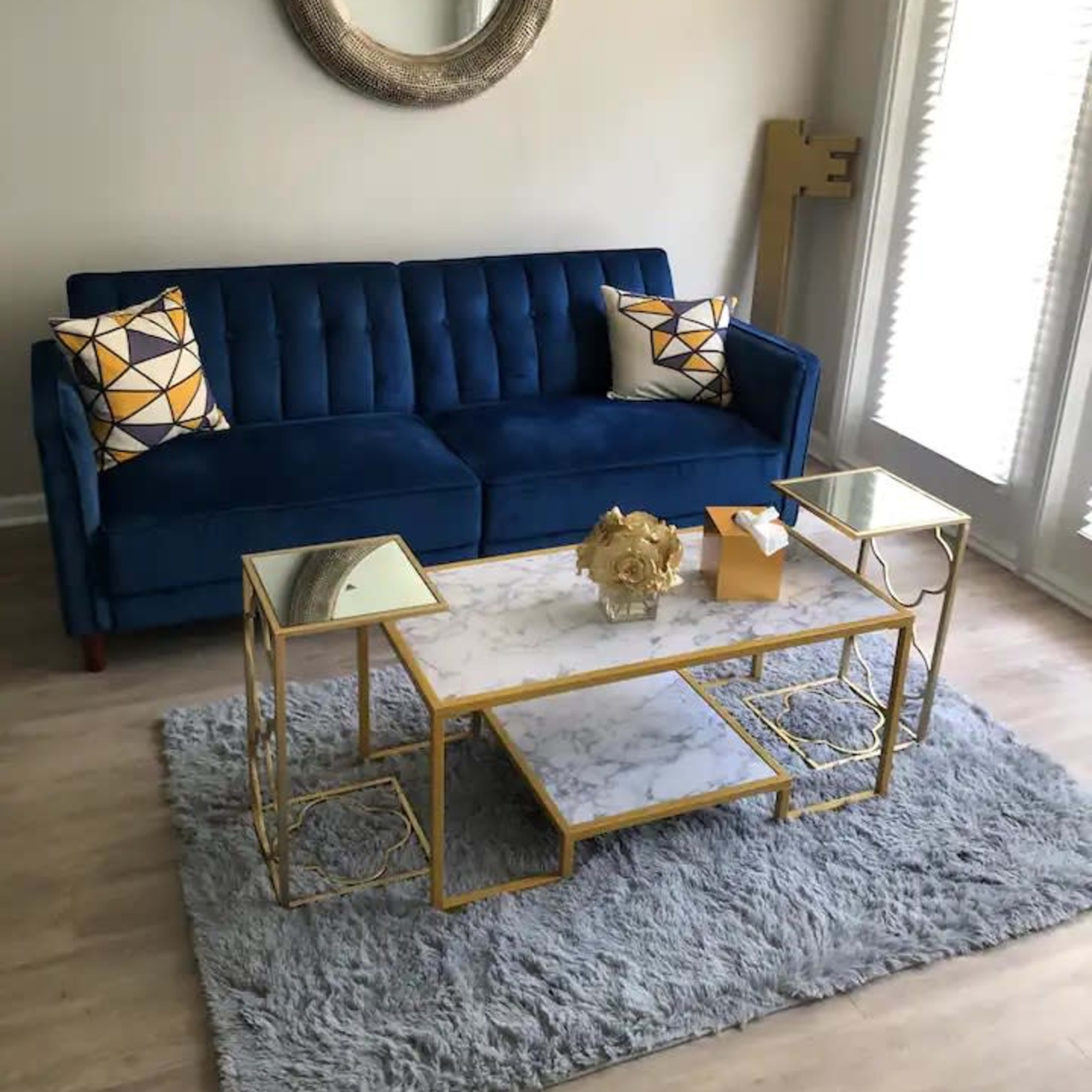 A modern living room with a blue velvet sofa, decorative pillows, a marble-top coffee table with gold accents, a light gray rug, and large windows allowing natural light to enter.