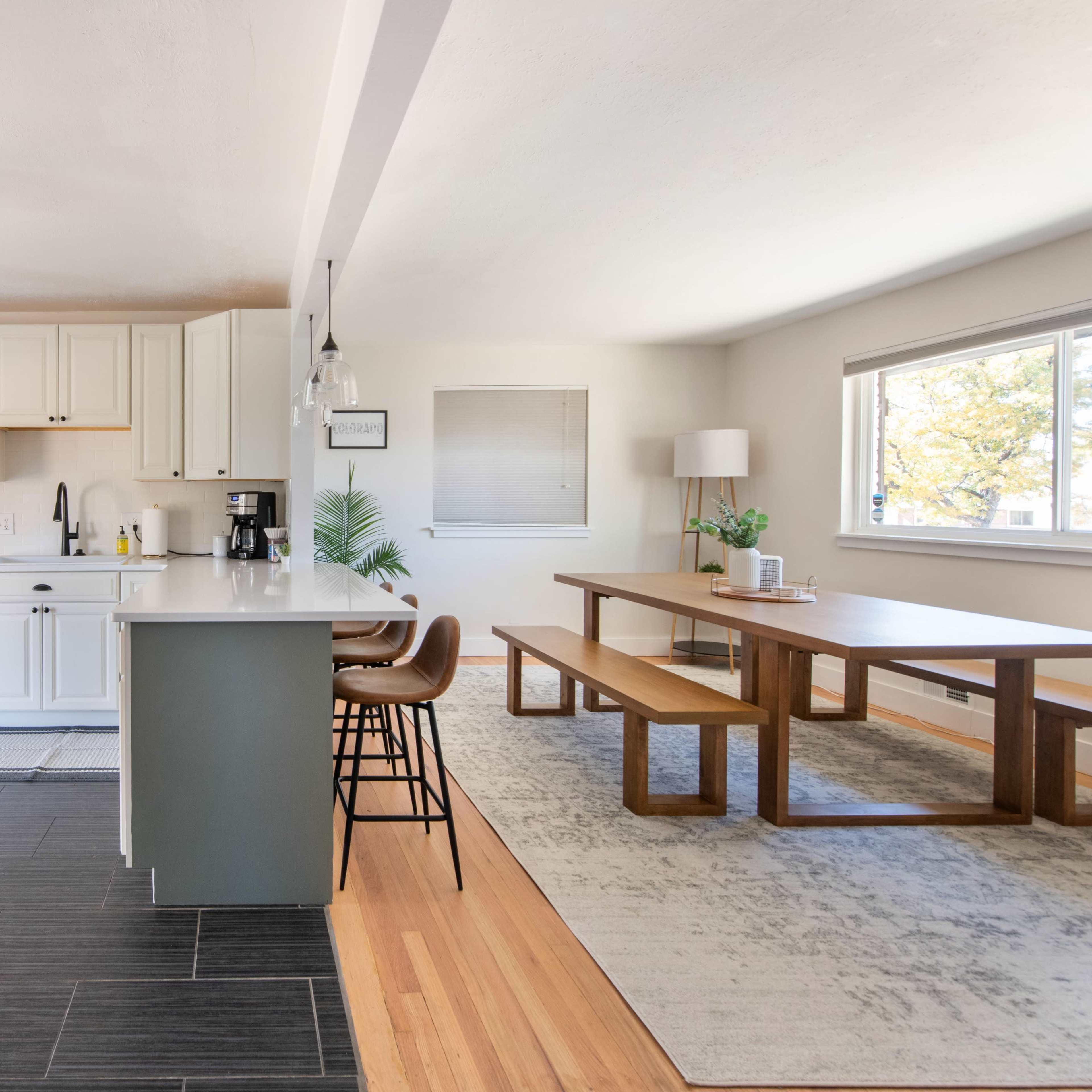 A modern kitchen transitions into a dining area with a wooden table and bench beside large windows.