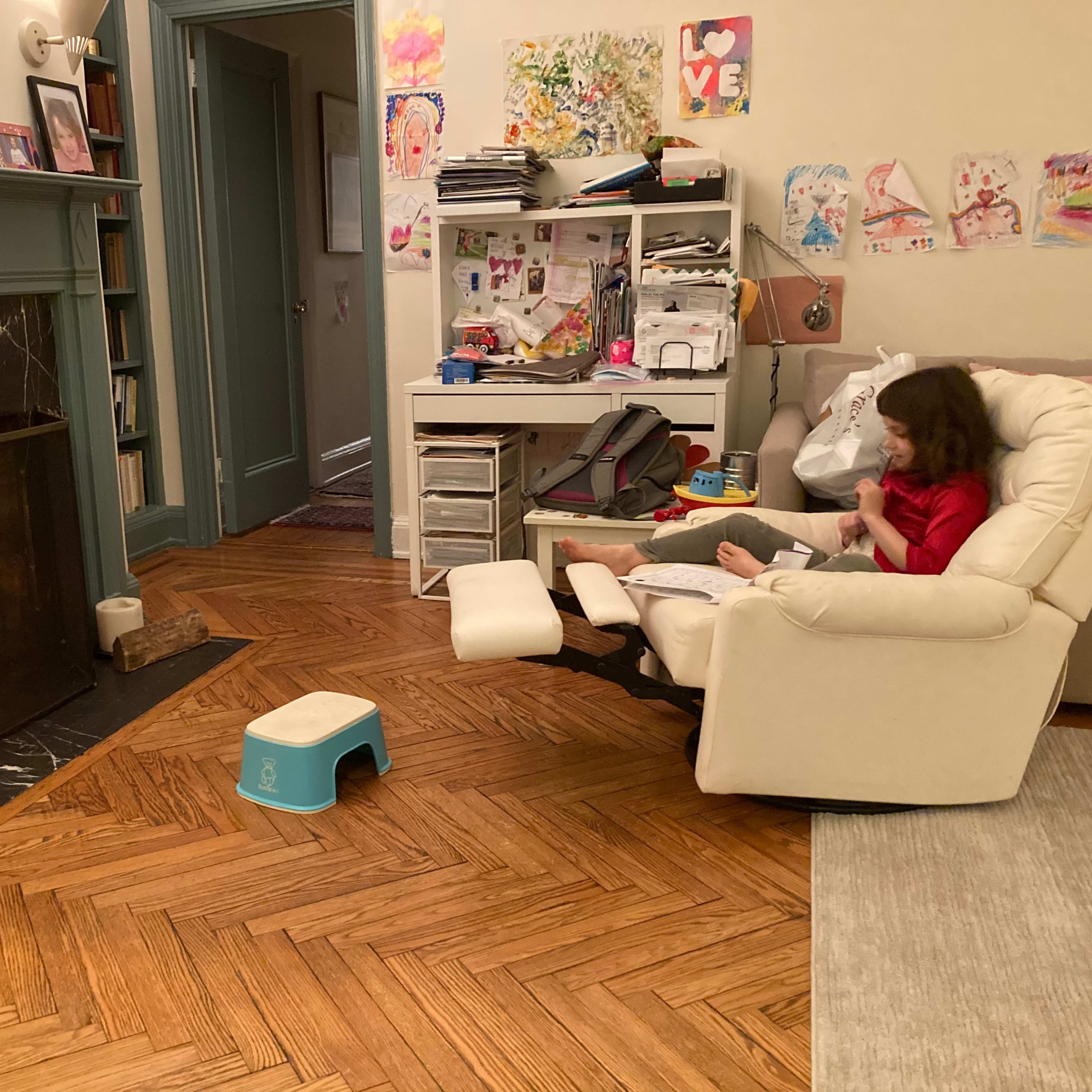 A child sits in a reclining chair near a fireplace, while a cluttered desk and colorful artwork decorate the room.