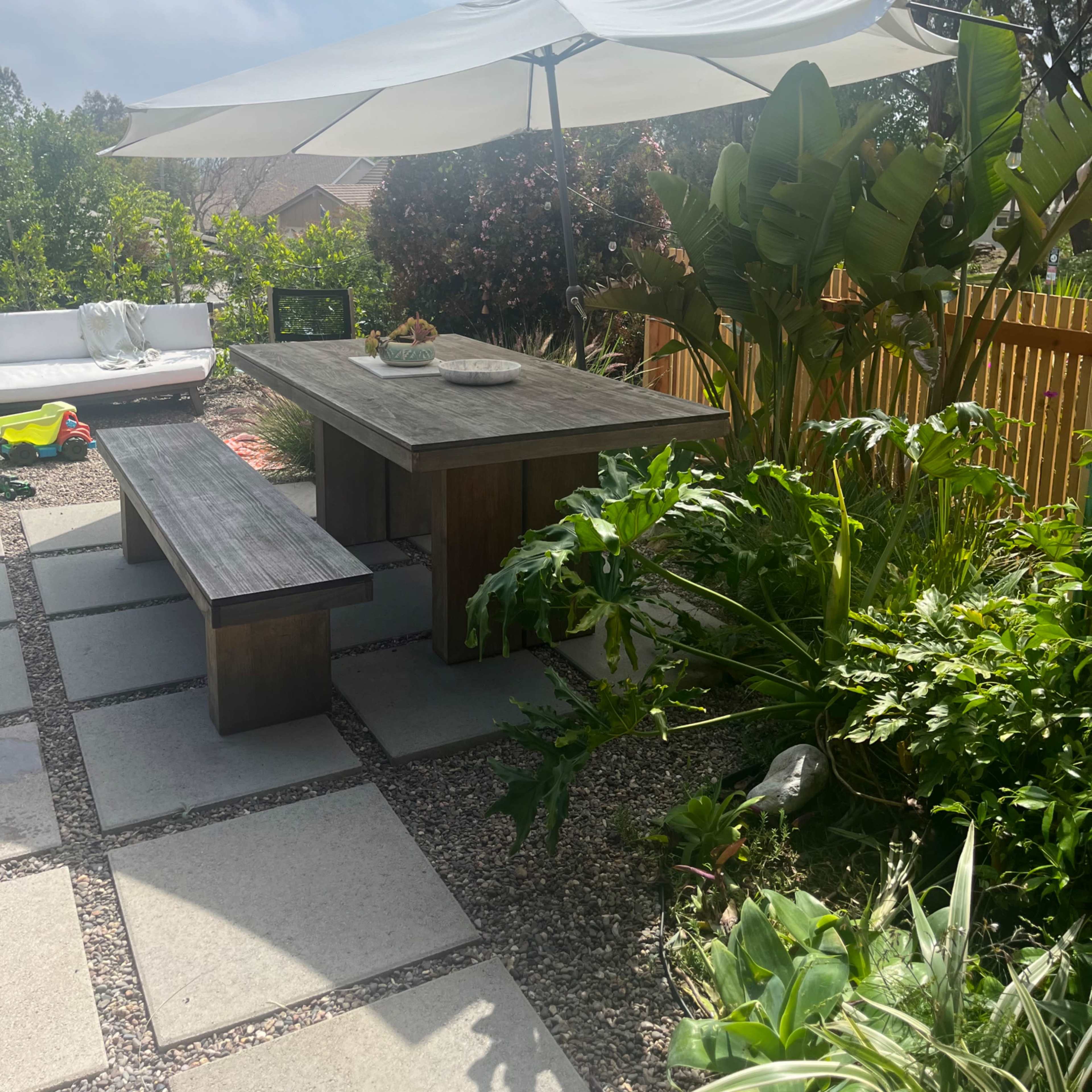 A wooden picnic table with a large umbrella is surrounded by vibrant greenery and pavers in a patio setting.