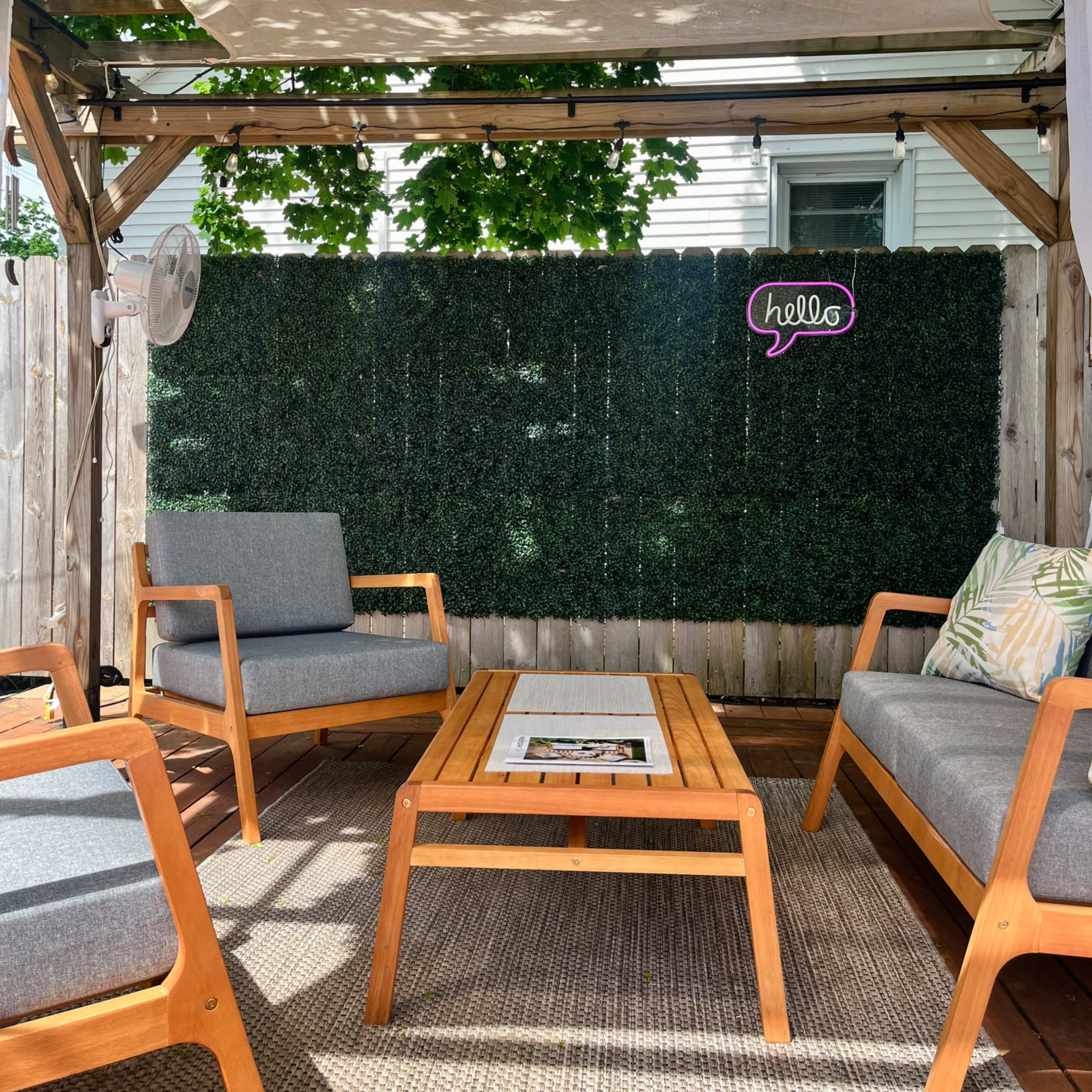 A shaded outdoor seating area with two wooden chairs and a coffee table, surrounded by a greenery backdrop and a "hello" sign on the wall.