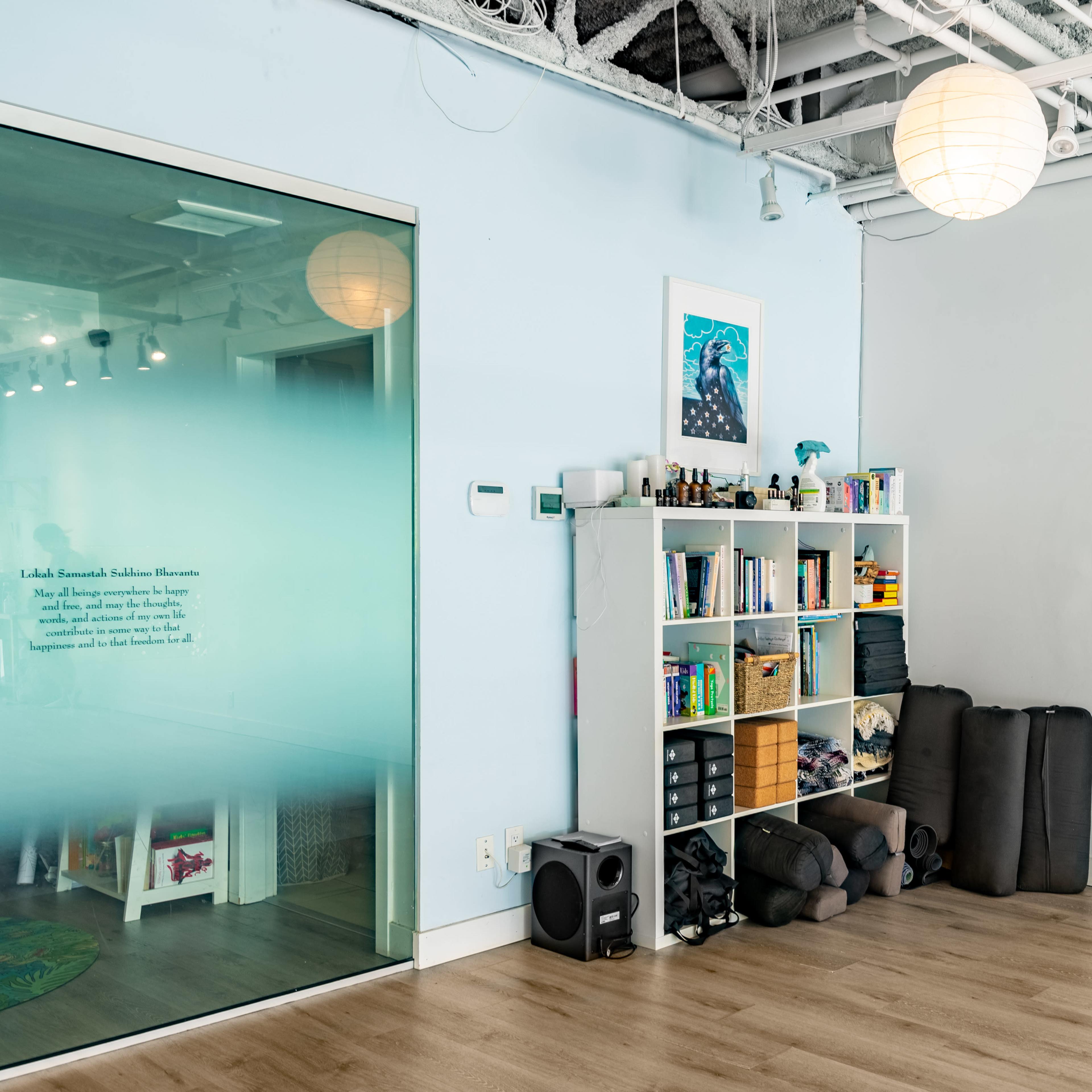 A tidy room features a shelving unit filled with books and various items, alongside neatly stacked exercise mats against a white wall.