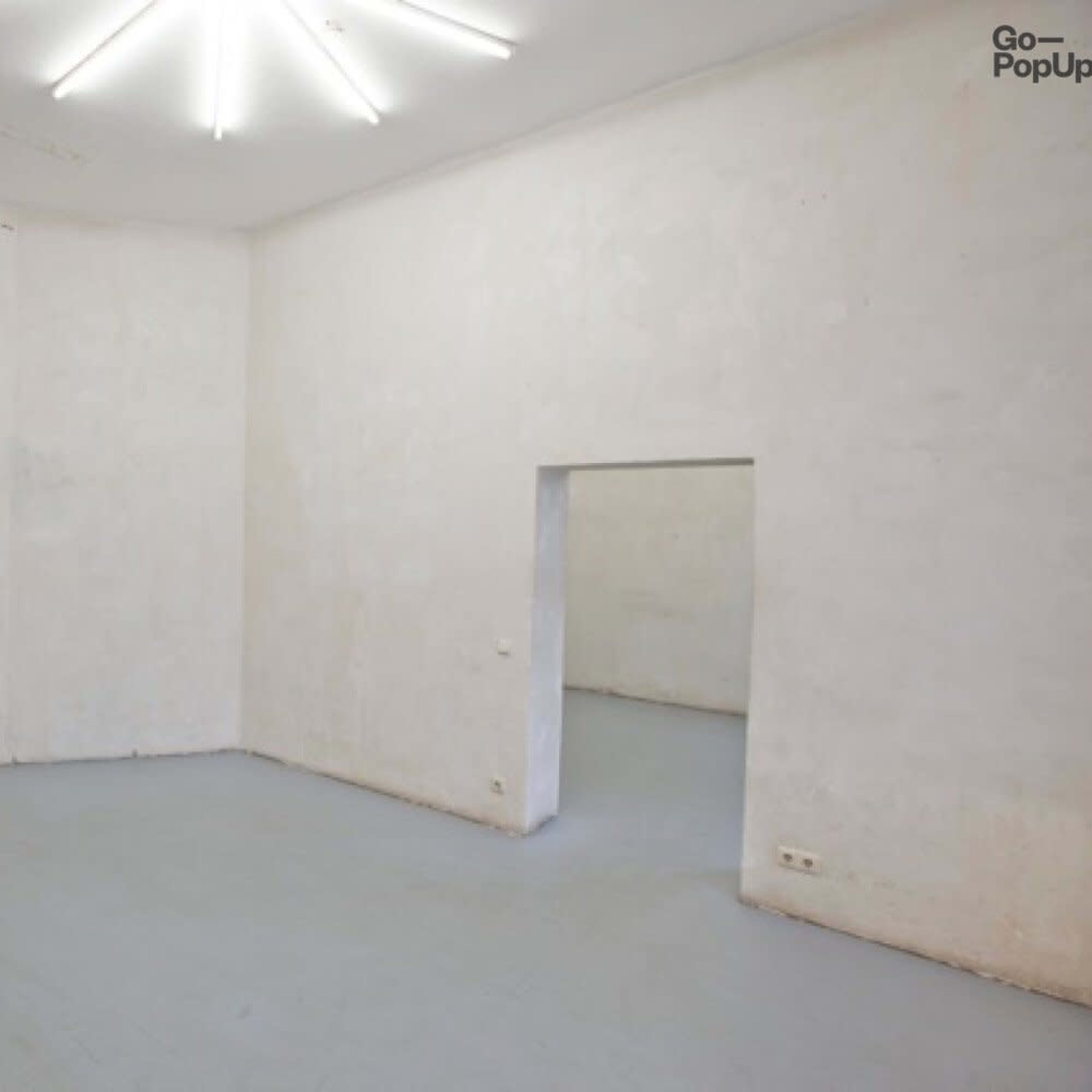 An empty, well-lit room with light gray flooring and plain, white walls, featuring doorways leading to adjacent spaces.