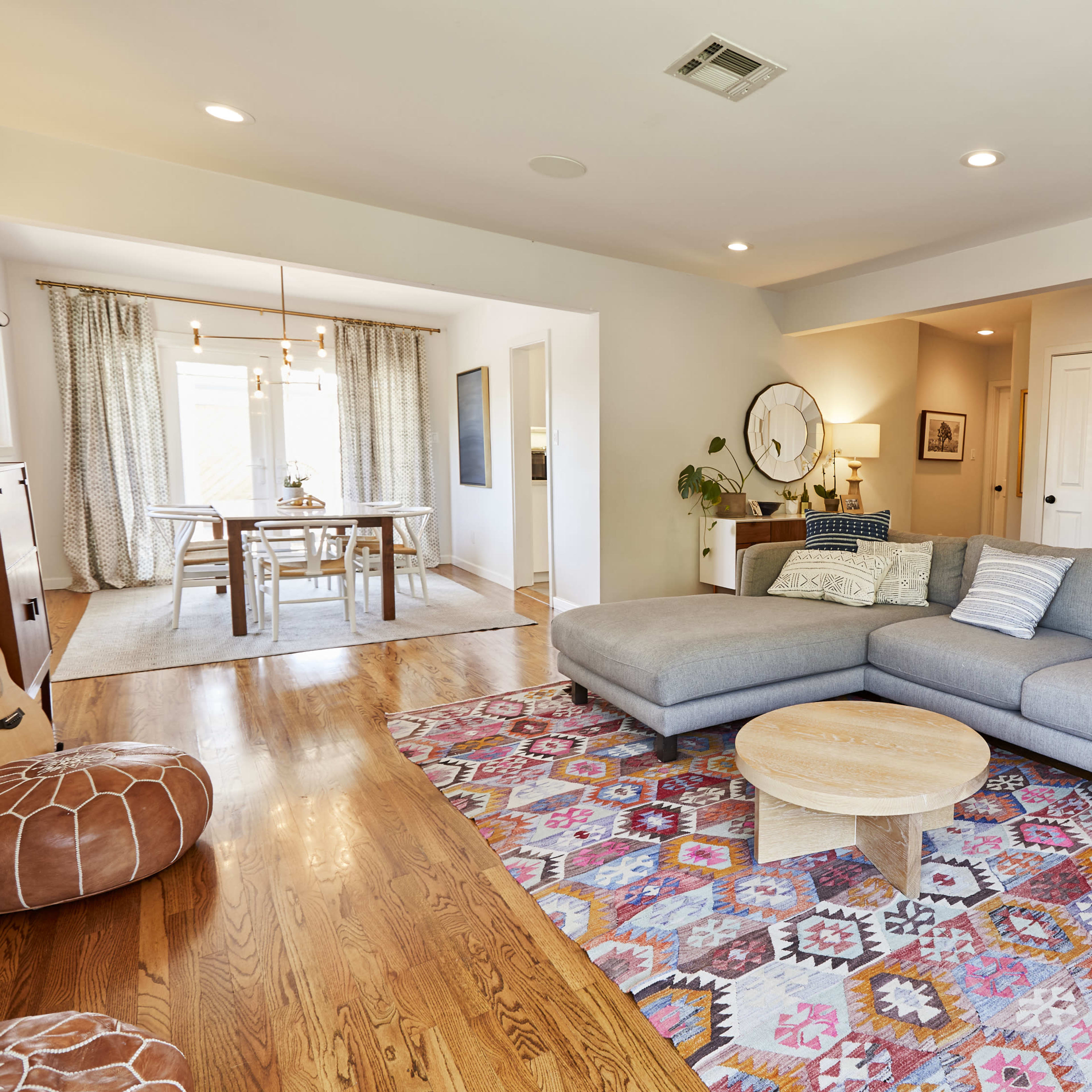 The living room features a gray sectional sofa, a round wooden coffee table, and a colorful area rug on hardwood floors, with an adjoining dining area visible in the background.
