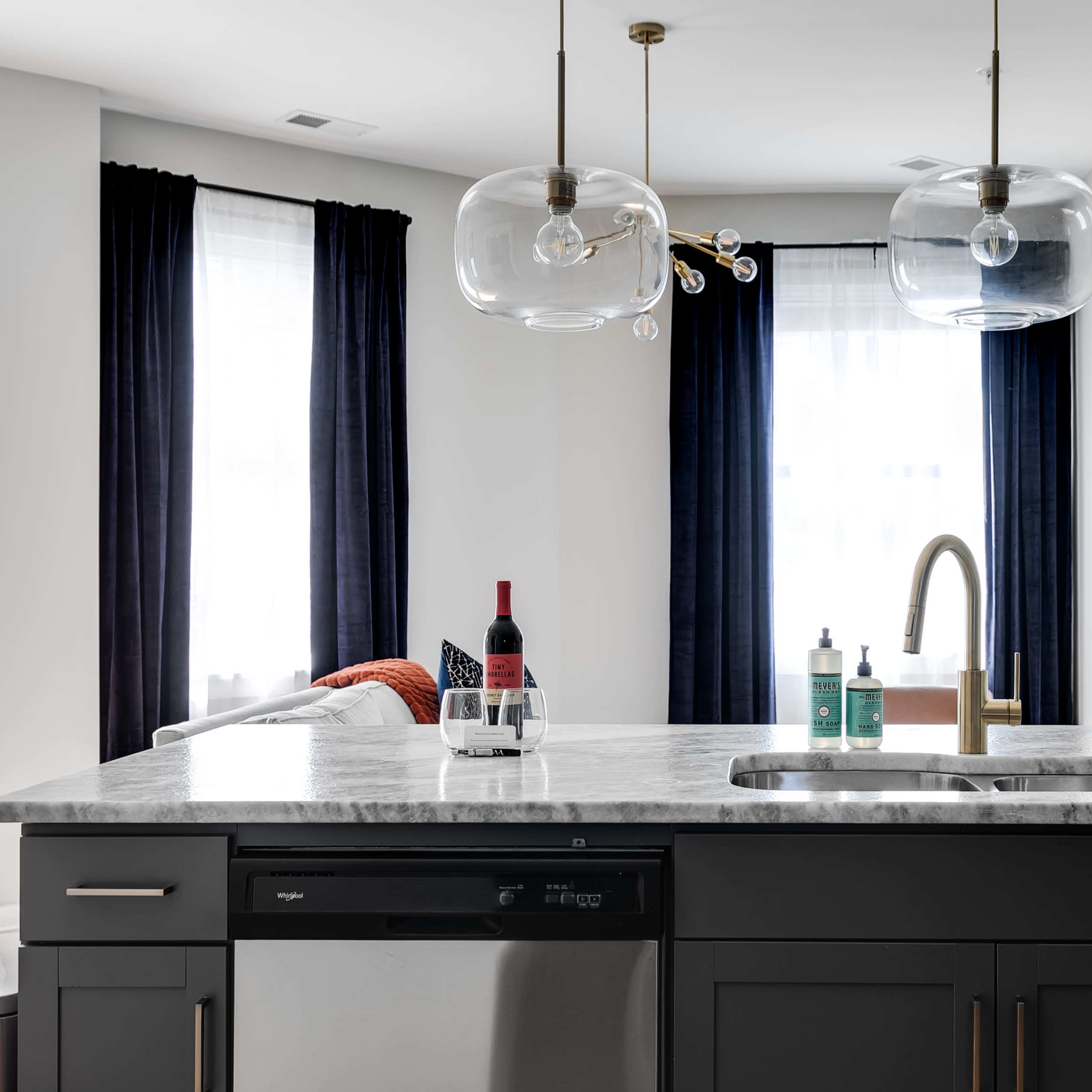A modern kitchen features a marble countertop, dark cabinetry, and pendant lights above the sink, with bottles and a wine glass on the surface.