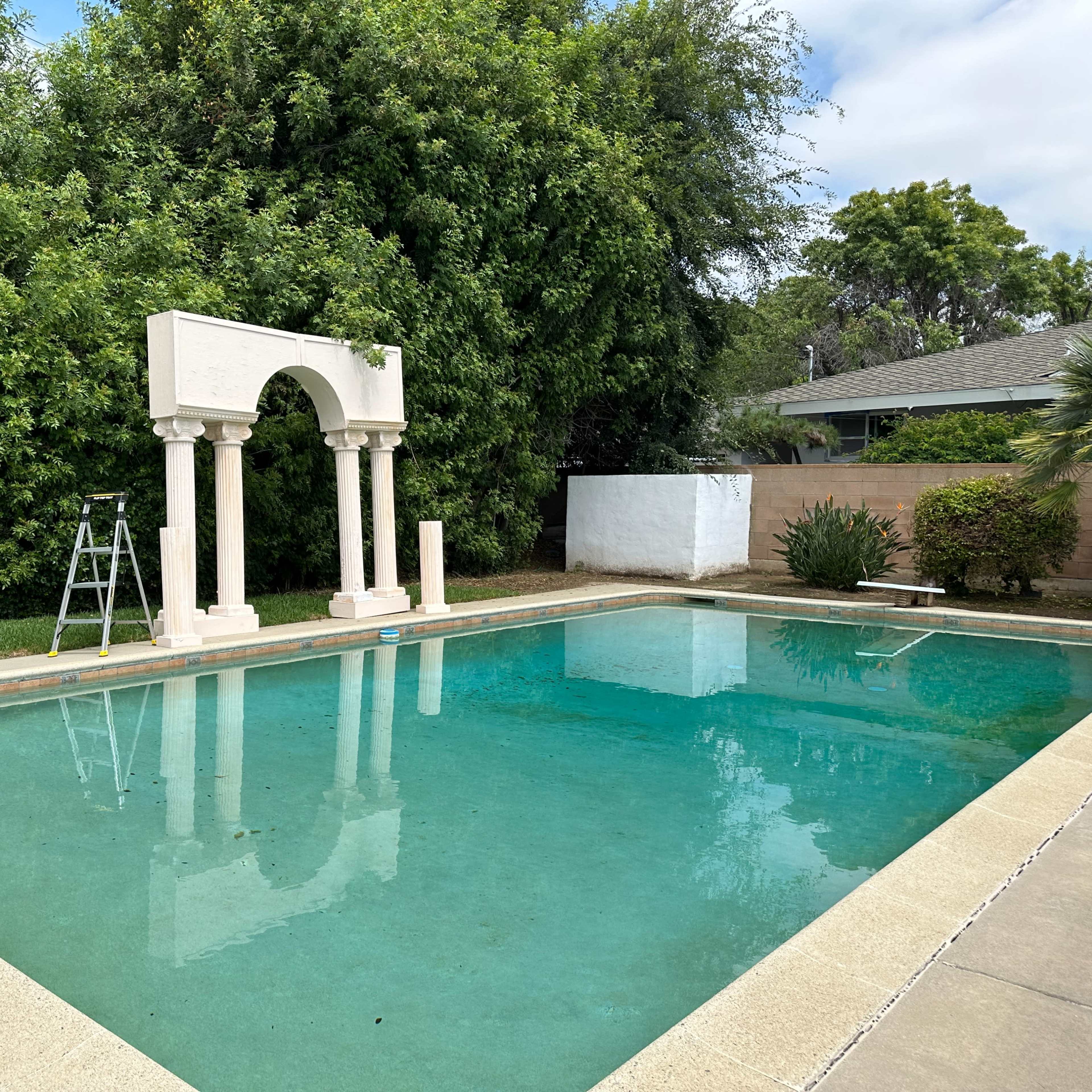 A rectangular swimming pool is surrounded by greenery and features a decorative arch at one end, with a ladder positioned nearby.