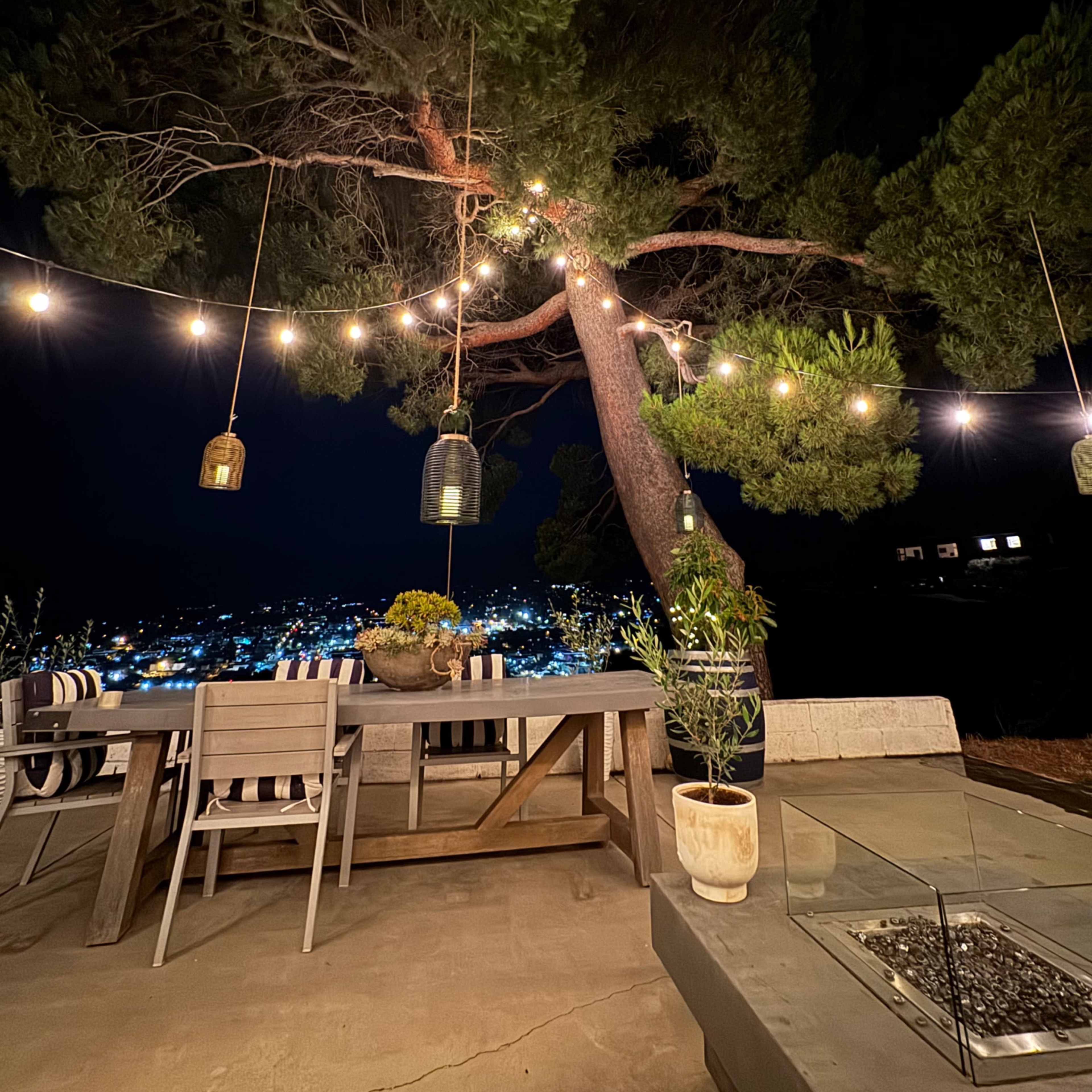 A patio table with chairs is set under a large tree adorned with hanging lights, overlooking a city illuminated at night.
