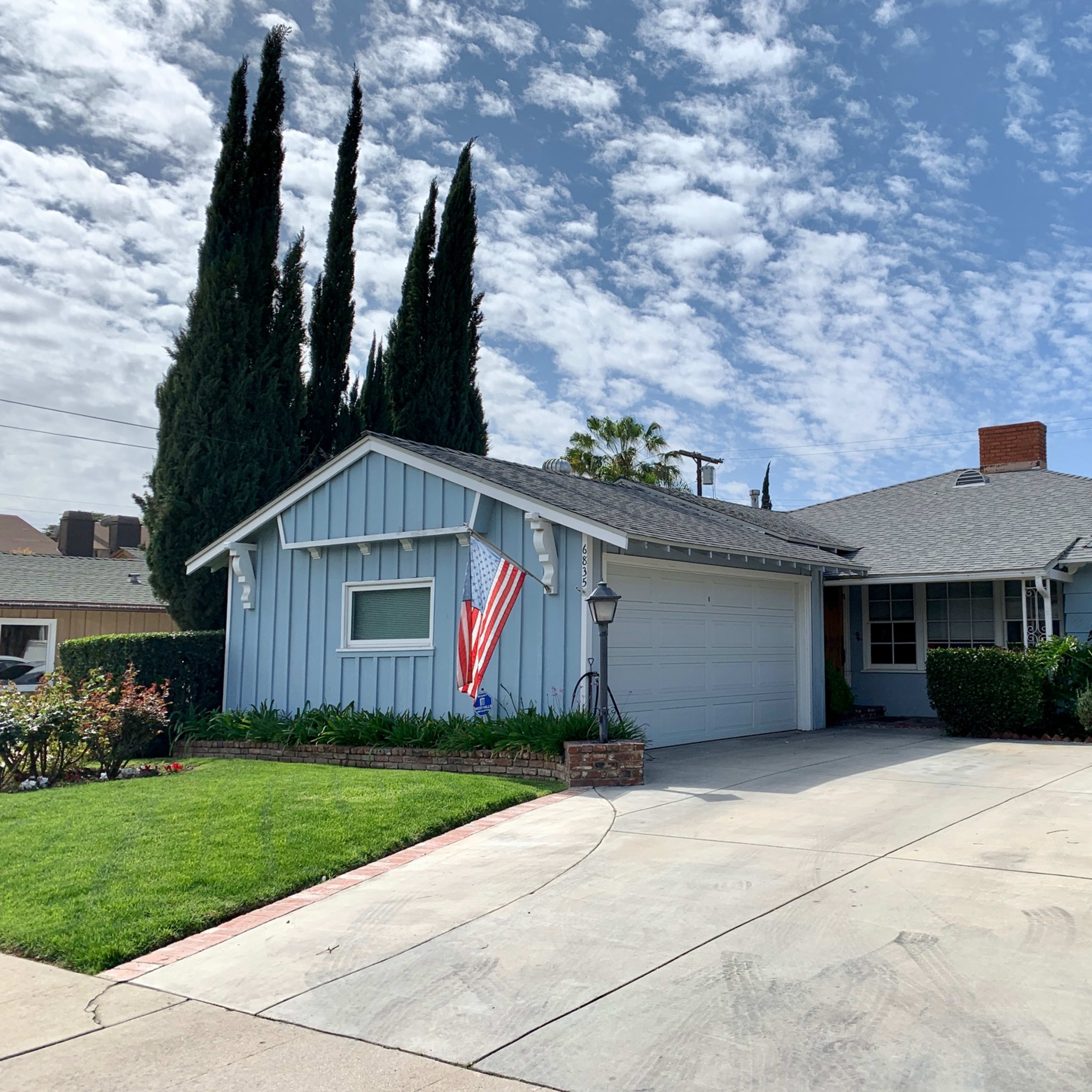 A single-story blue house with an American flag and a well-maintained front yard is positioned on a residential street.