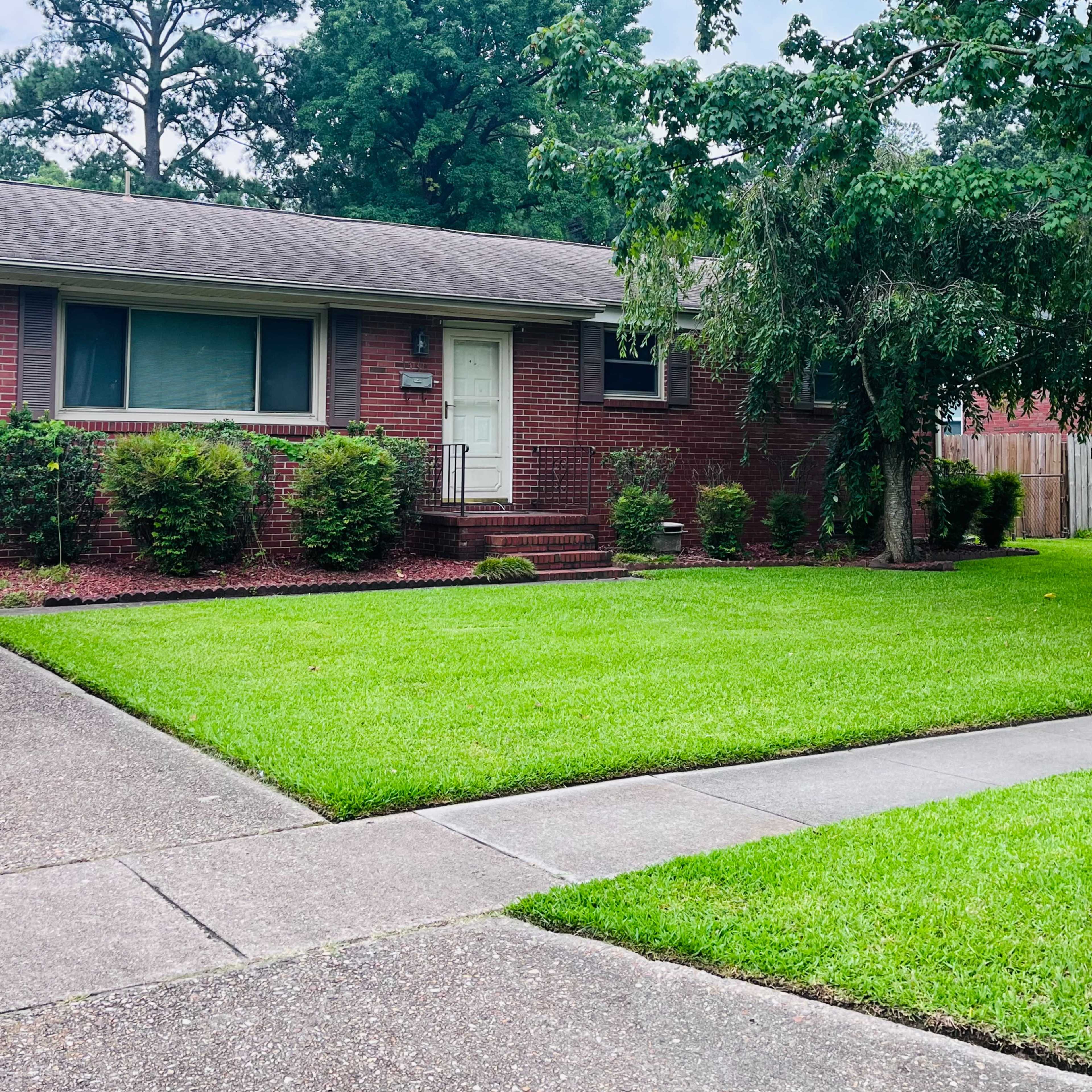 A red brick house with a white door is situated on a well-maintained lawn, surrounded by shrubs and trees.