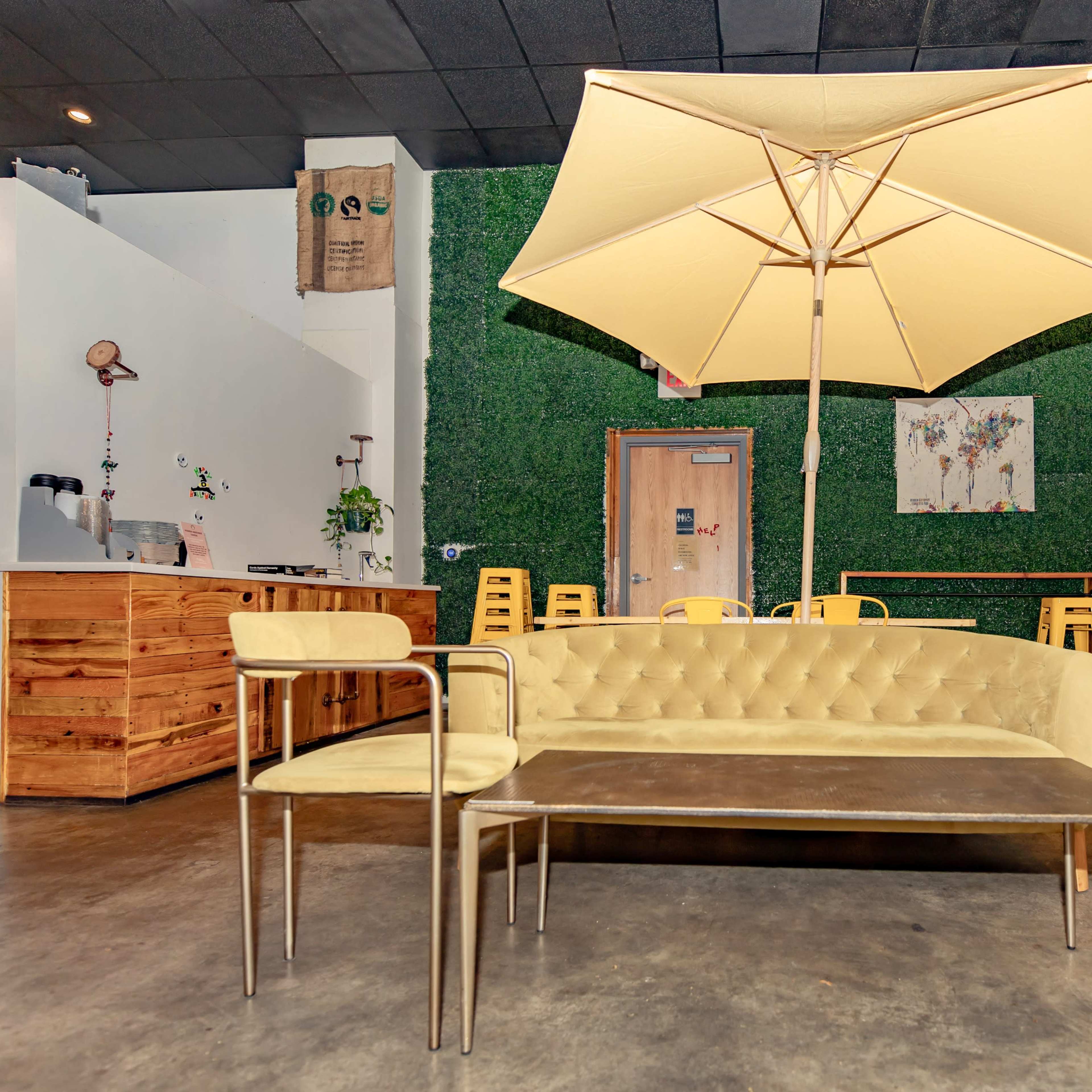 A modern interior featuring a yellow tufted sofa, a metal coffee table, and a large umbrella, with green wall decor and wooden furniture in the background.