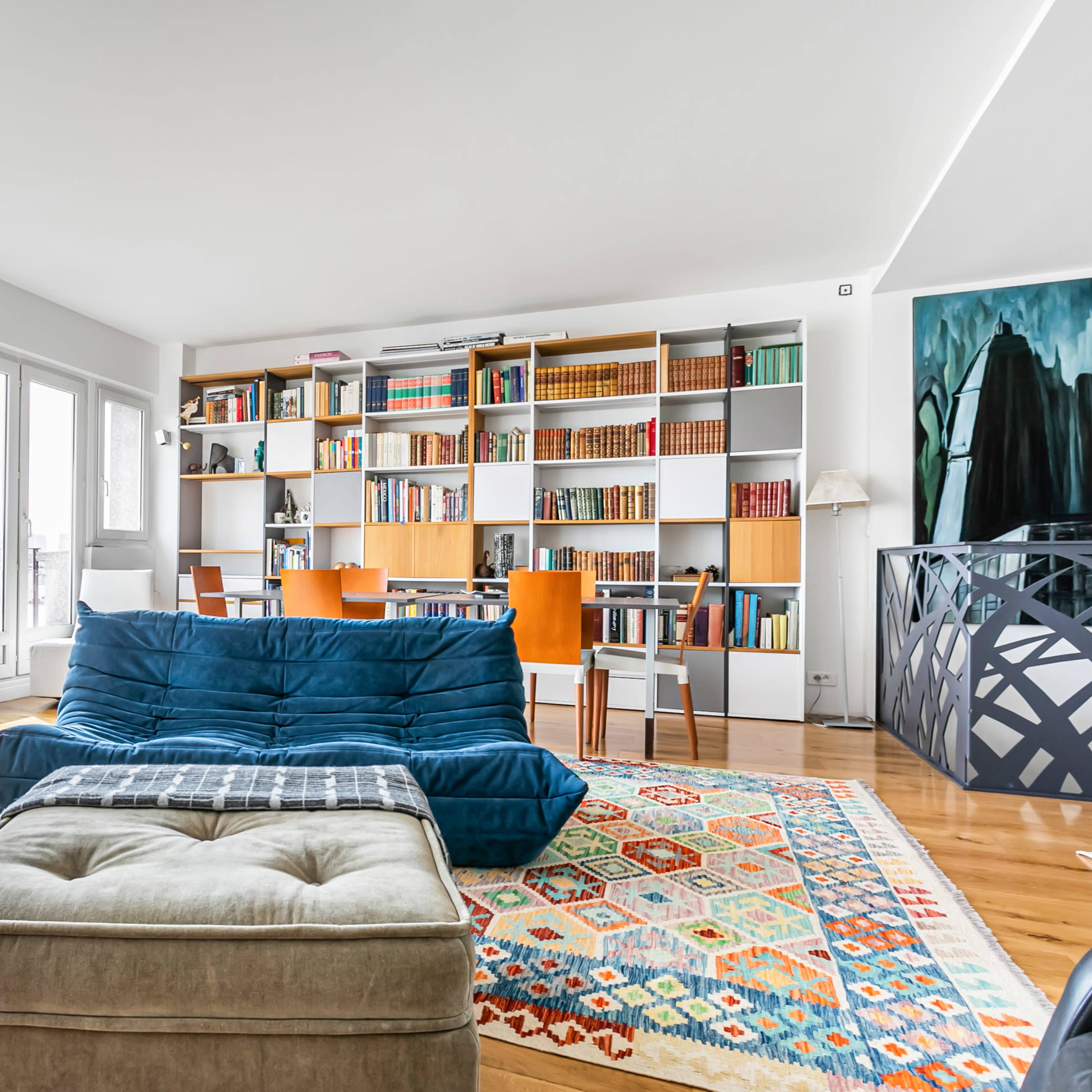 A contemporary living room with a blue sofa, a brown armchair, and a bookshelf filled with books in front of large windows.