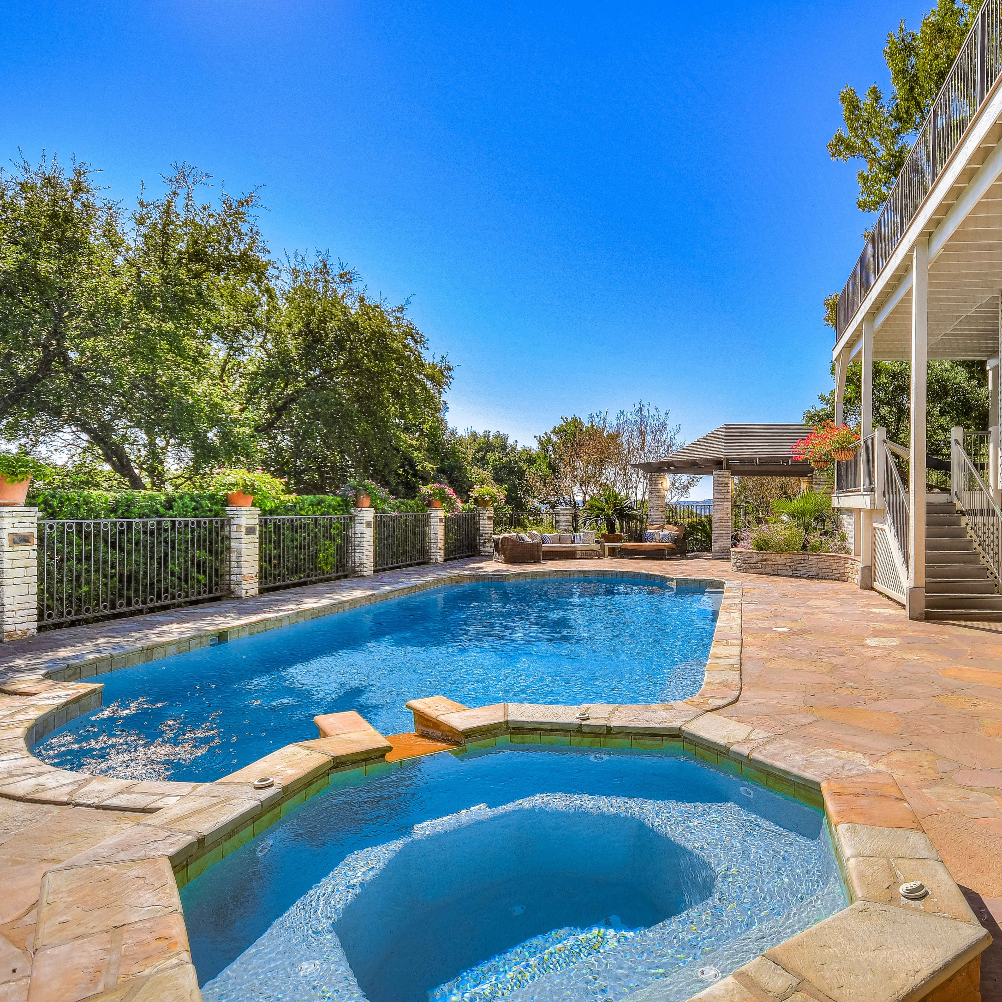 A backyard pool area with a blue pool, a spa section, stone patio, and a house with a covered porch.