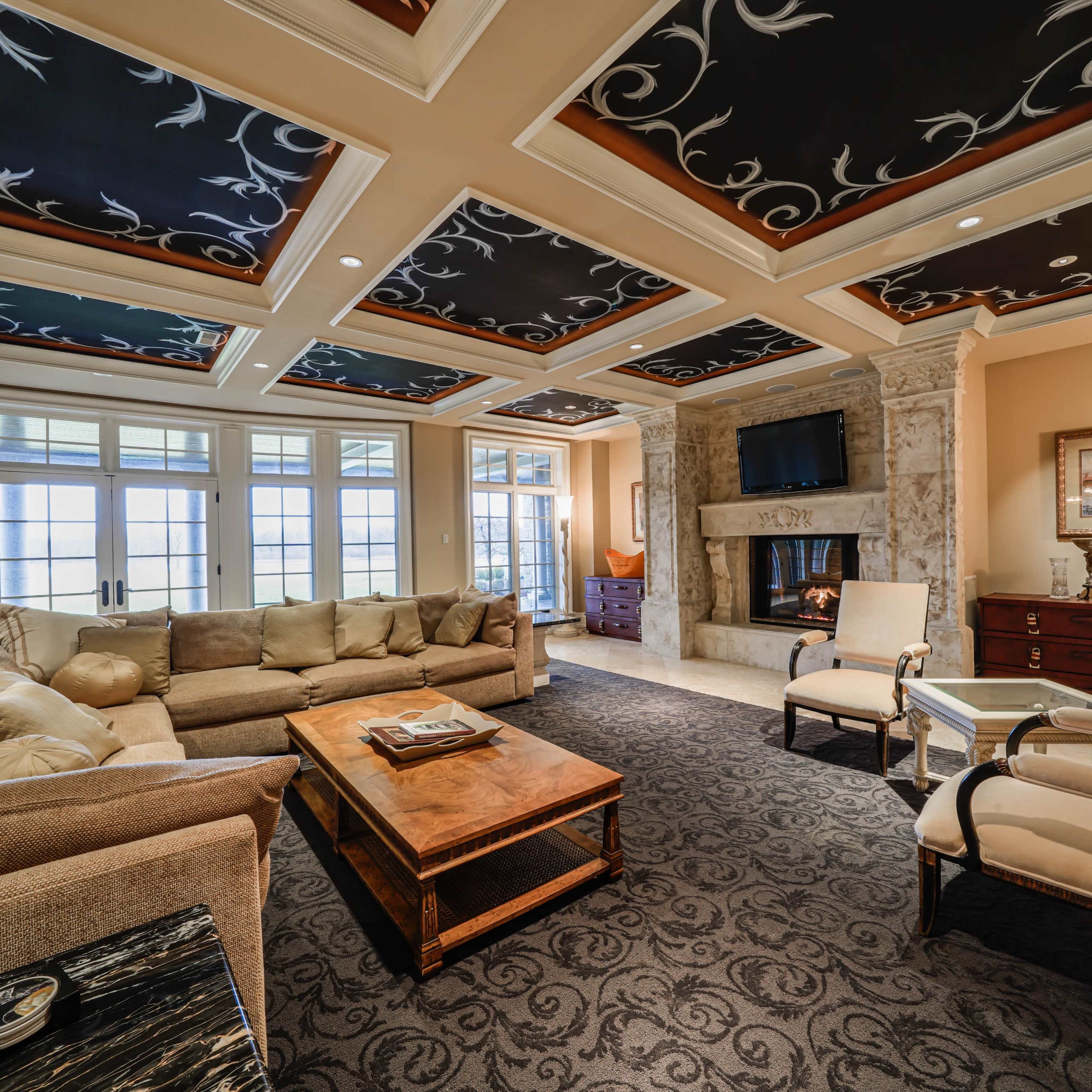 The living room features a large sectional sofa, a wooden coffee table, and a stone fireplace, with decorative ceilings and multiple windows overlooking a view.