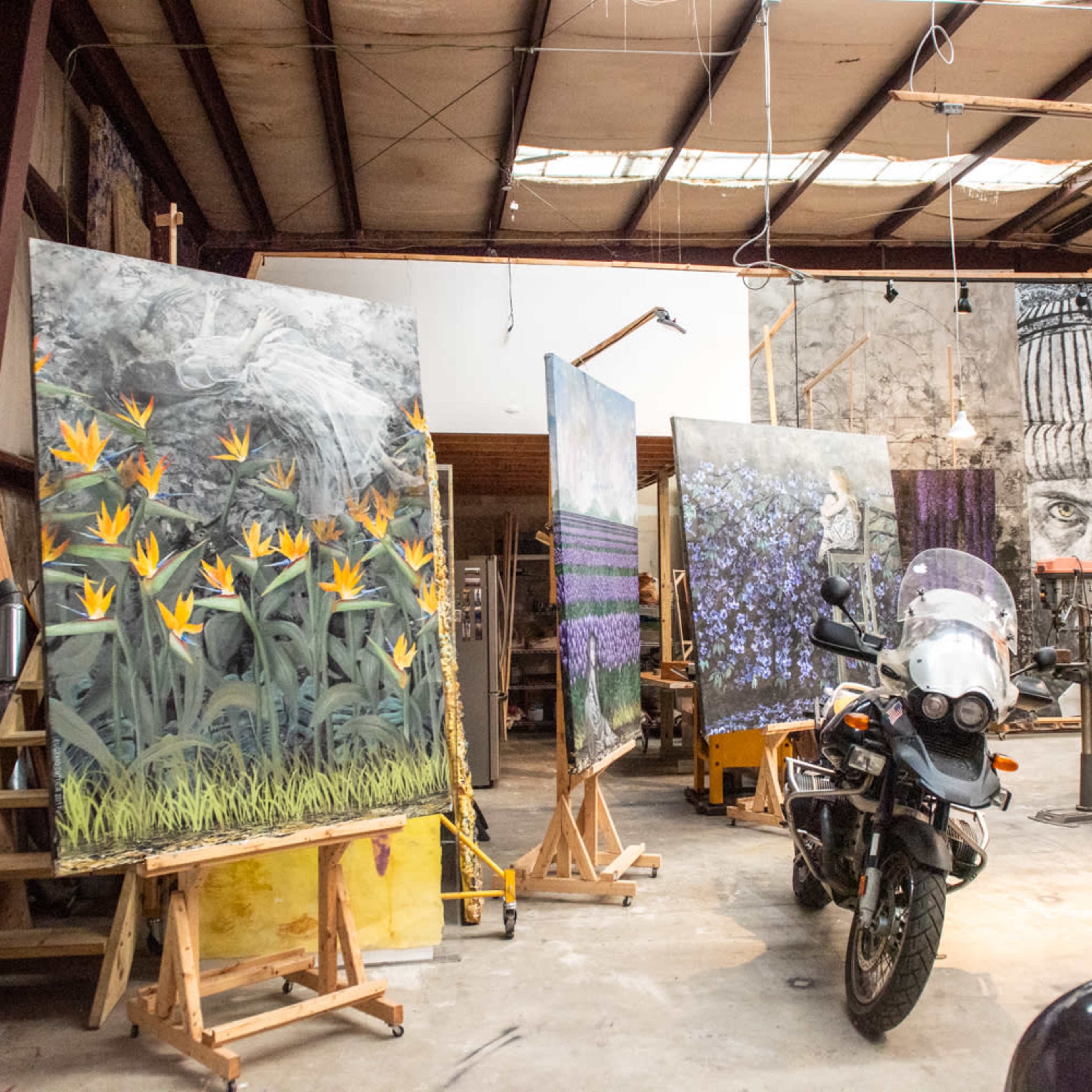 An art studio features large canvases on easels, displaying vibrant floral paintings, with a motorcycle parked in the foreground.