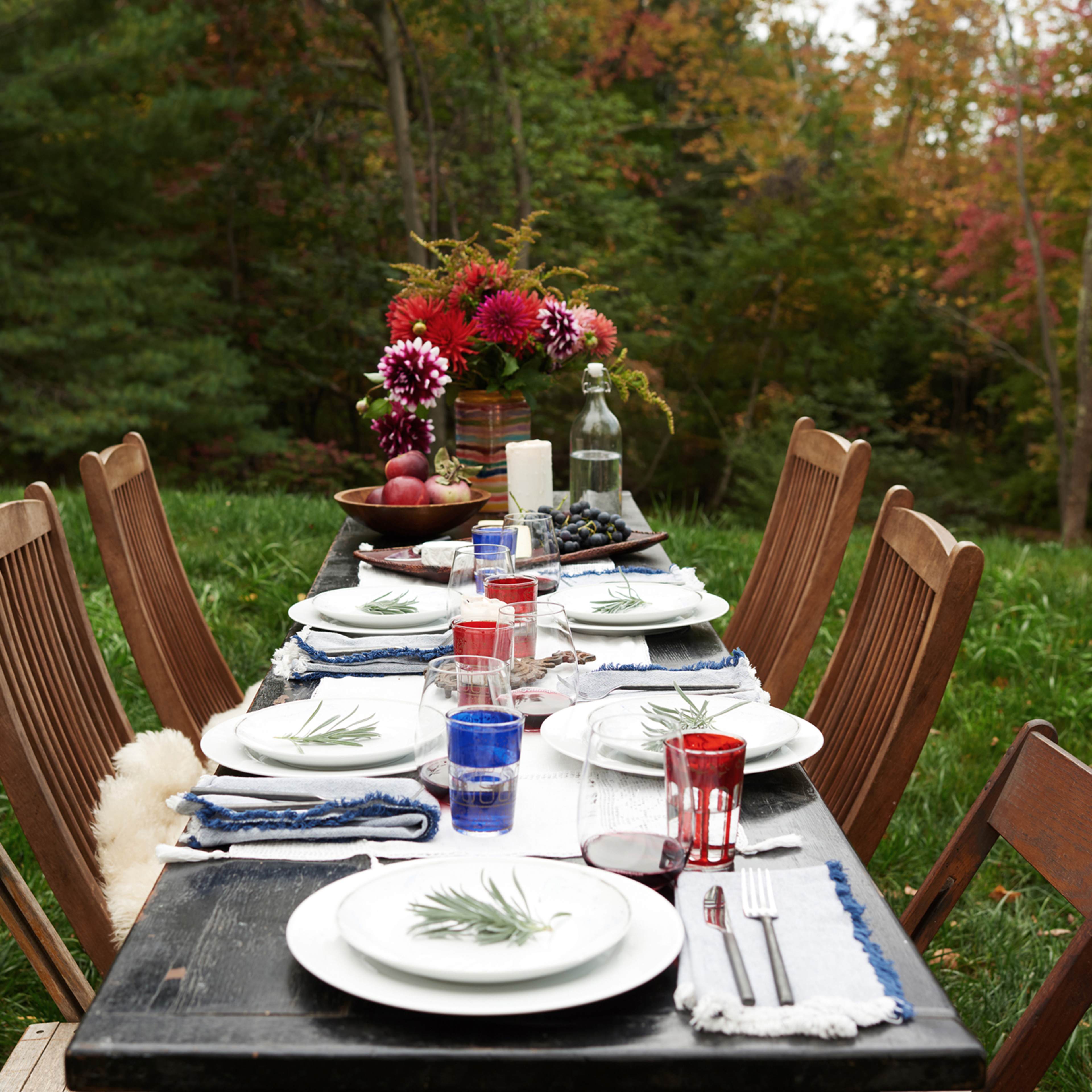 A long outdoor dining table is set with plates, glasses, and a vase of flowers, surrounded by greenery and autumn foliage.