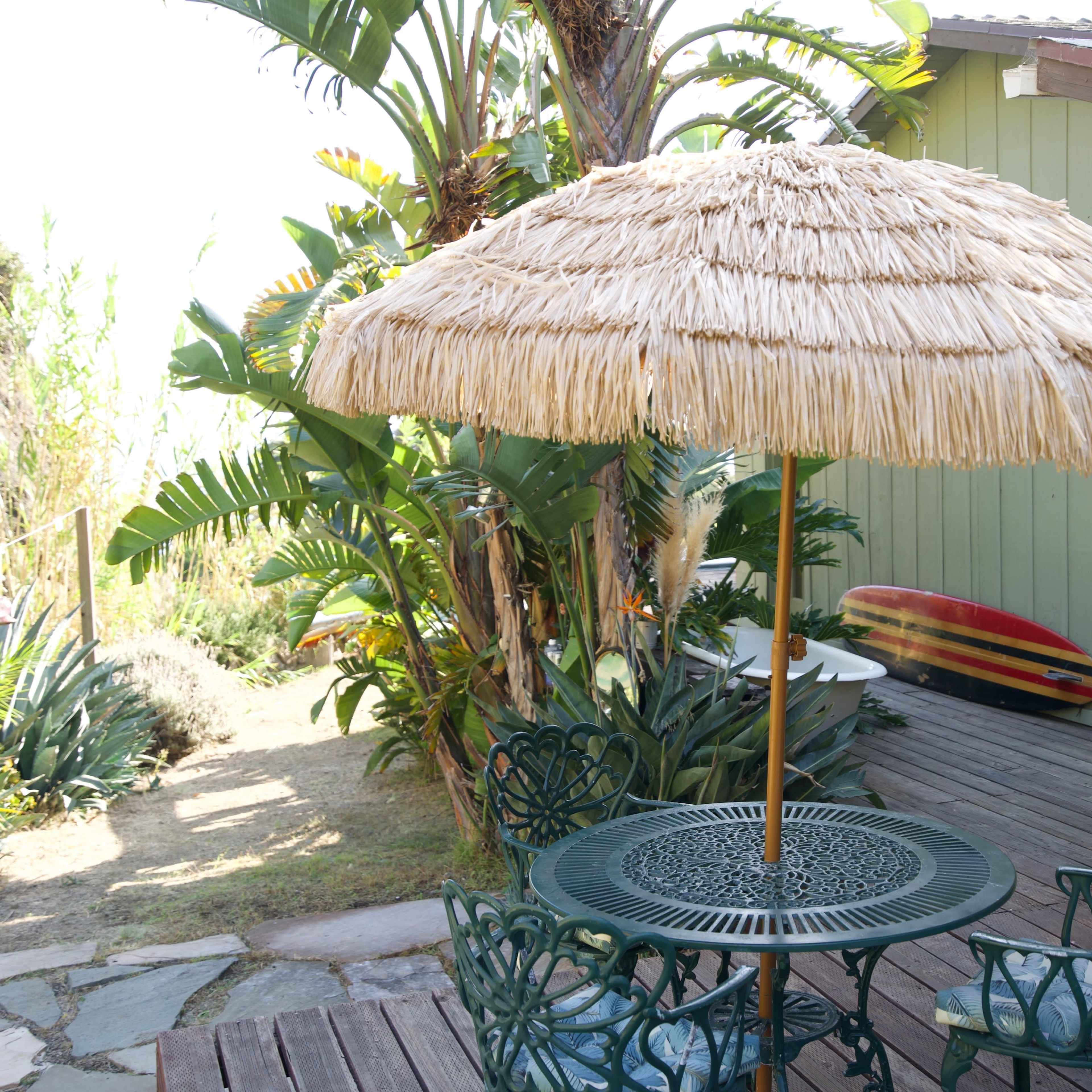 A straw umbrella stands over a metal table and chairs surrounded by tropical plants and a wooden deck near a building with green walls.