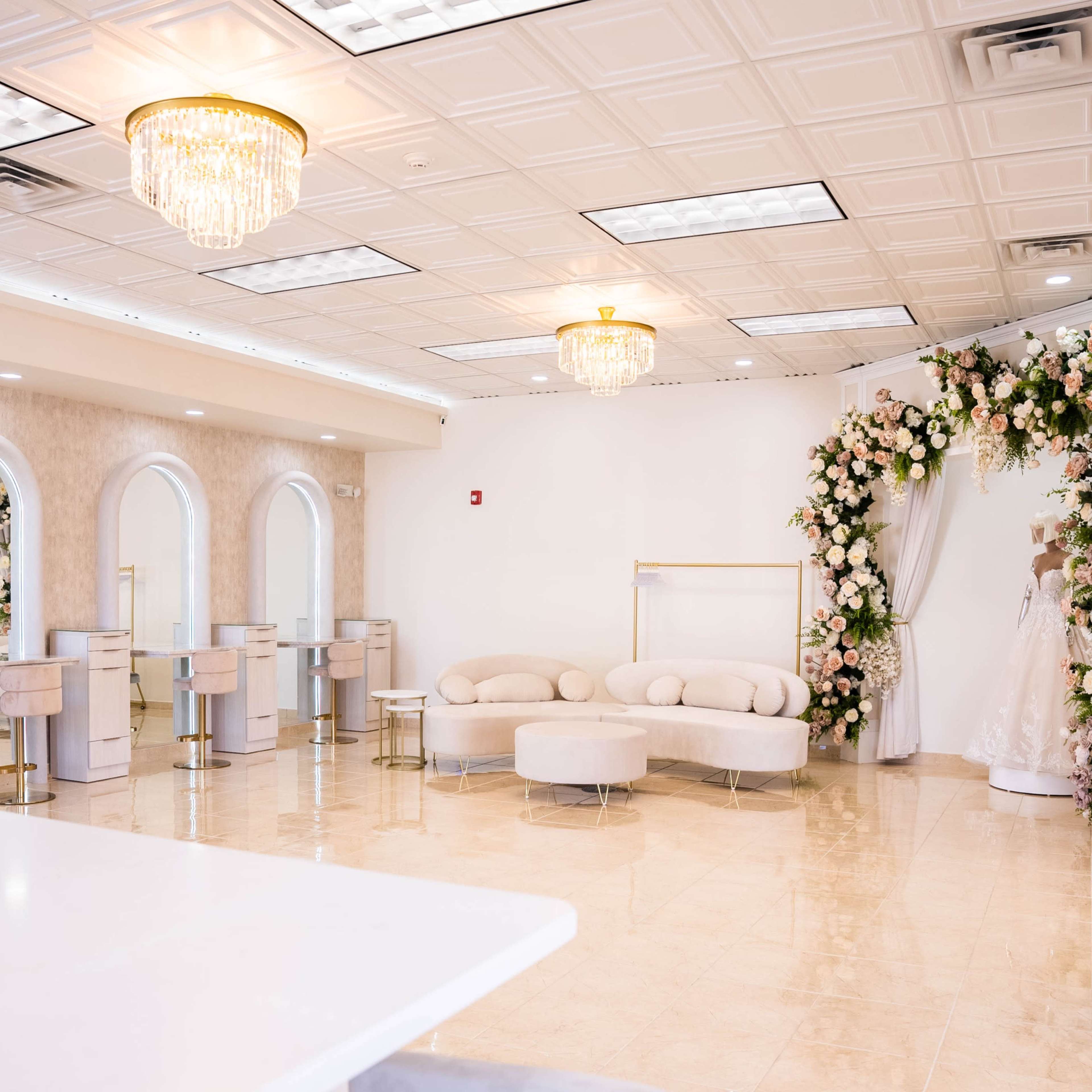 A brightly lit event space featuring elegant chandeliers, light-colored walls, a floral display, and seating arrangements with modern furniture.