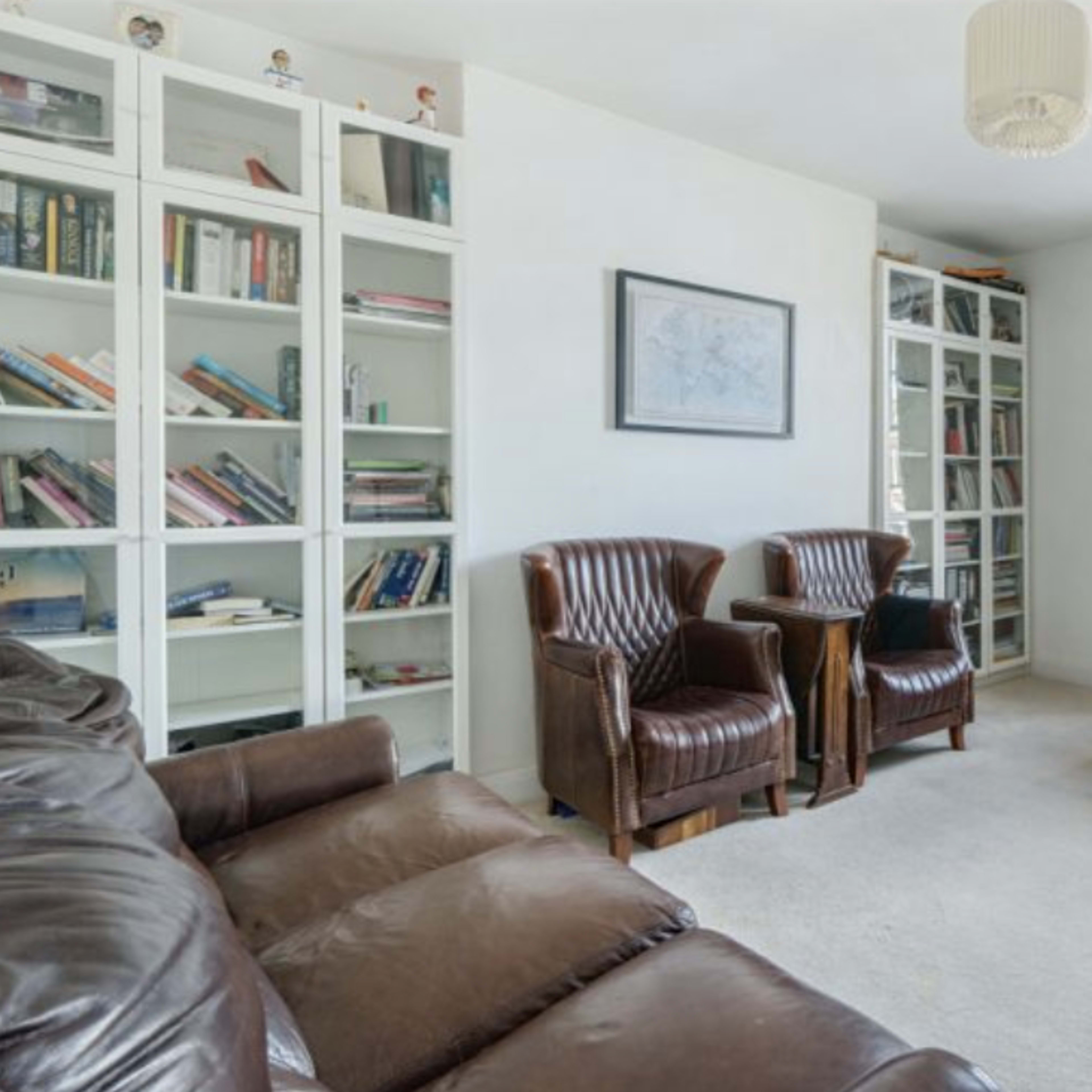 A well-lit home office with a desk, a computer, two leather armchairs, and bookshelves filled with books.