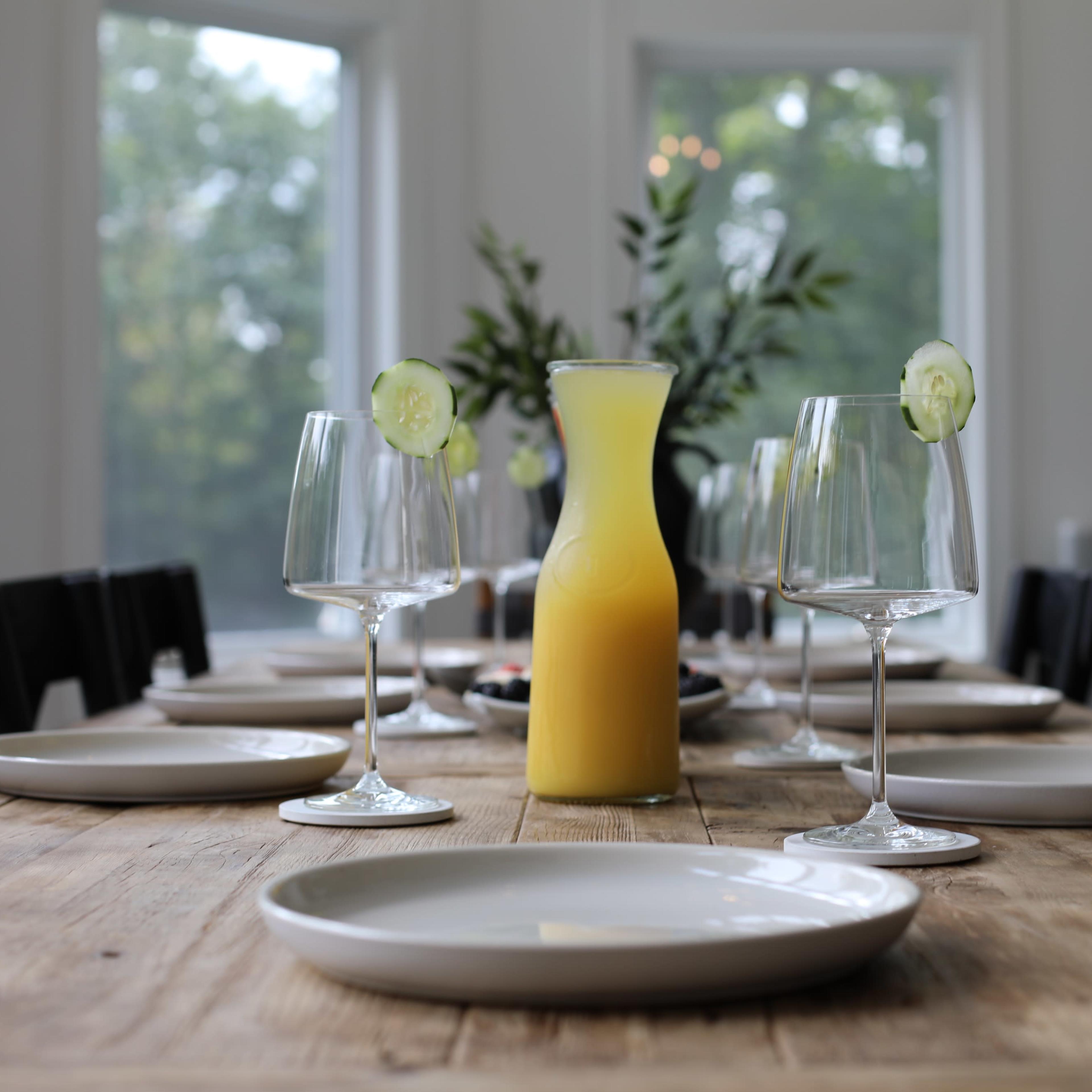 A wooden dining table is set with white plates and two glasses beside a pitcher of orange juice, accented by cucumber slices on the rim of the glasses.
