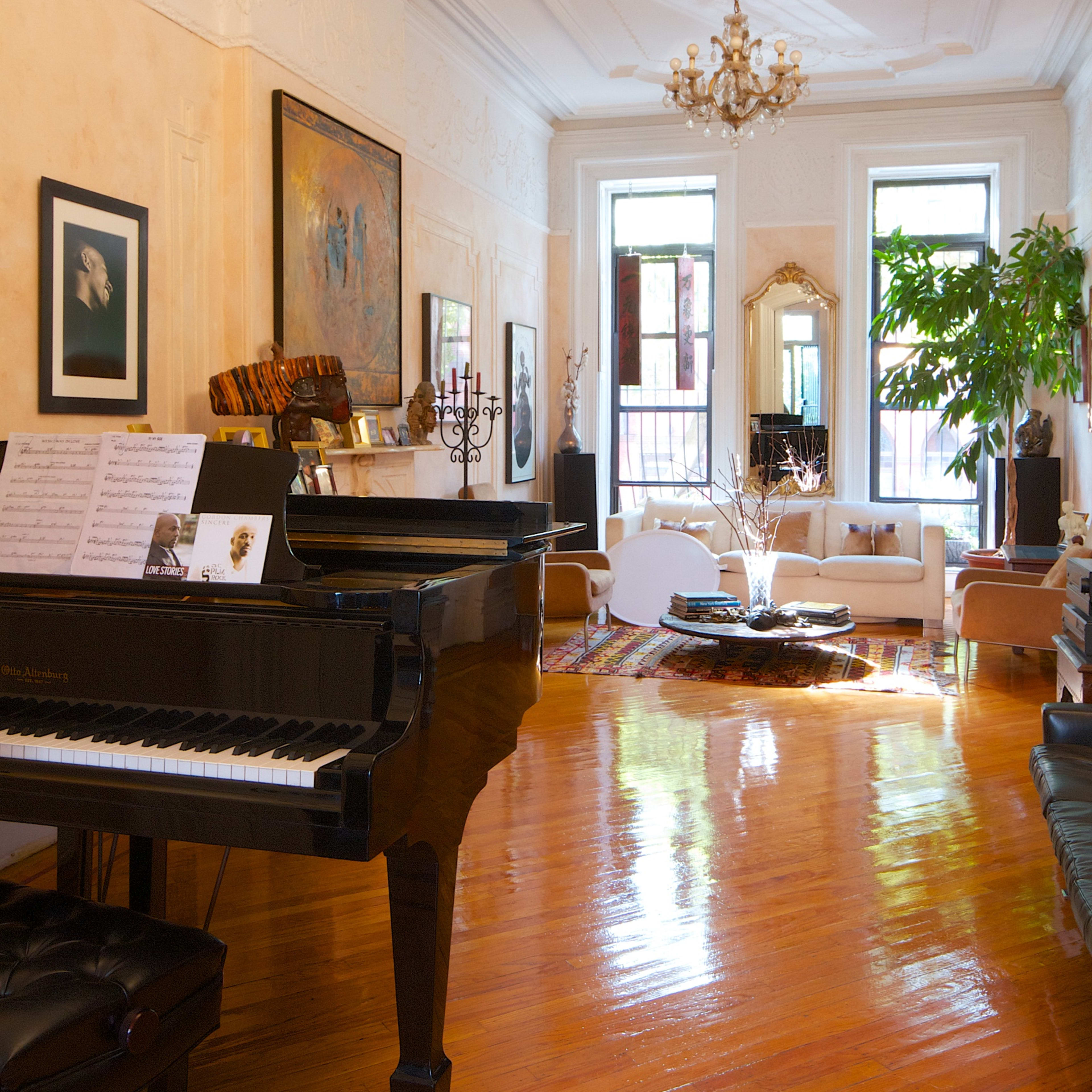 A spacious room with a grand piano on one side, a chandelier hanging from the ceiling, and a large window allowing sunlight to illuminate a seating area with a plant.