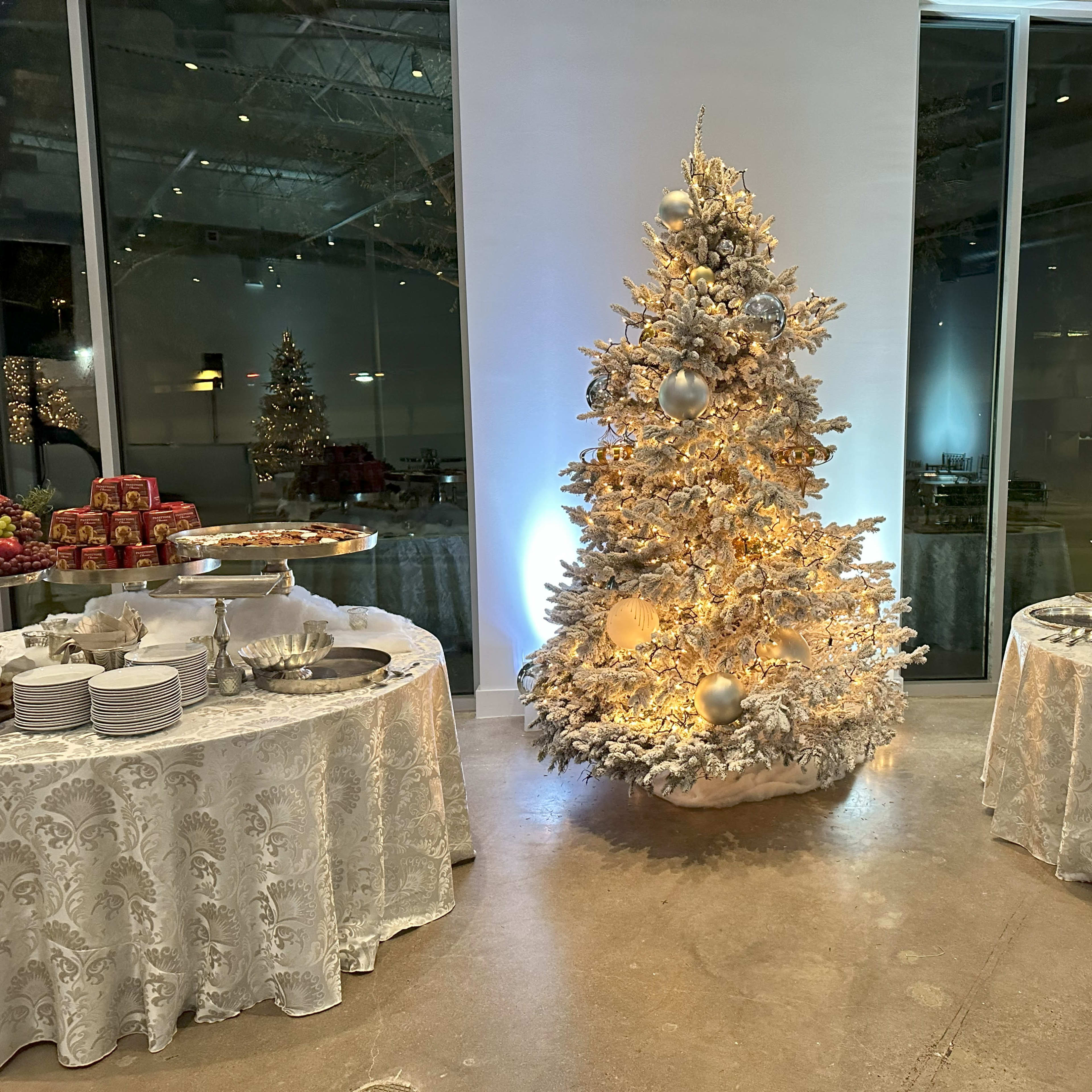 A decorated white Christmas tree with lights stands in the center of a room, flanked by two tables set with food and dishware.