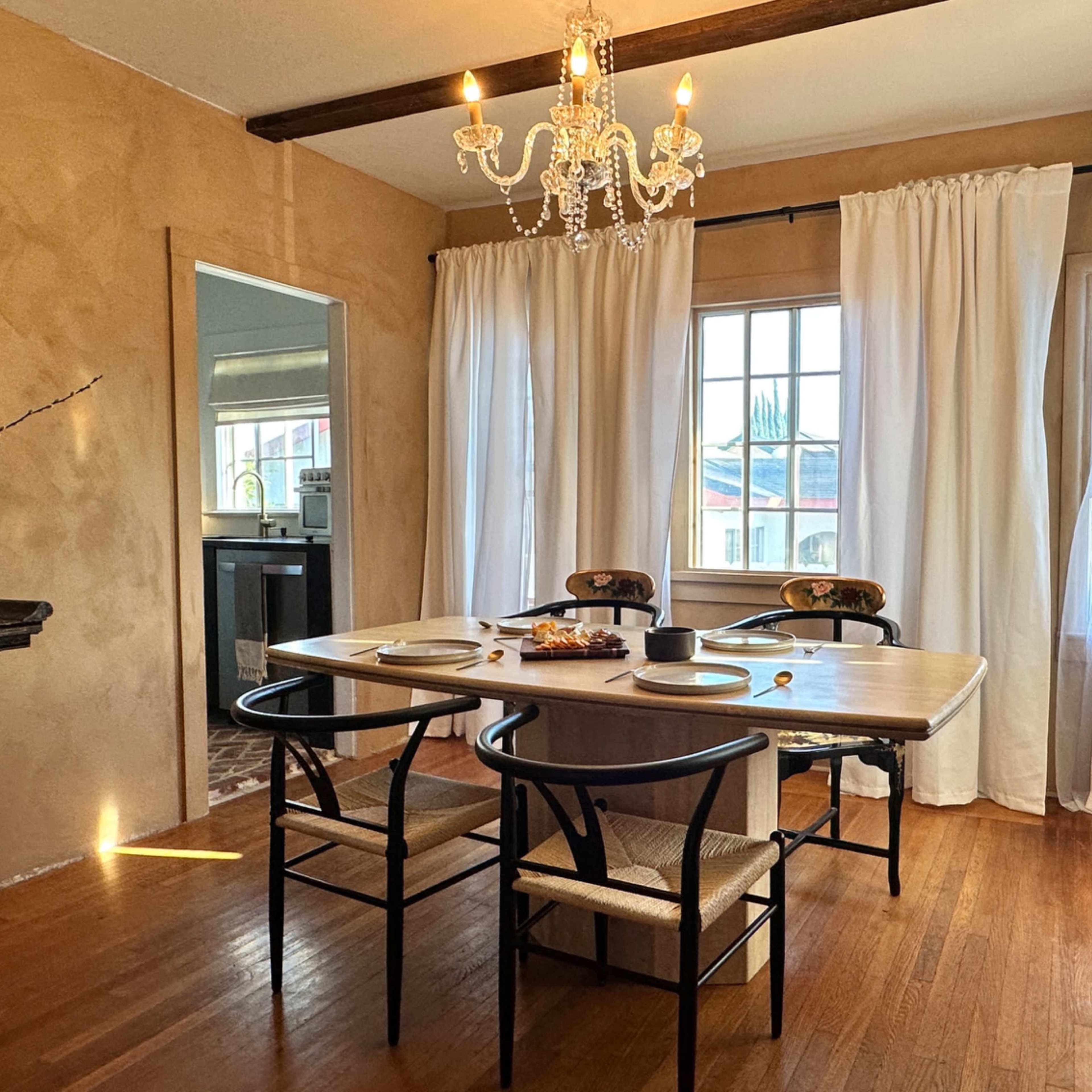 A dining room features a wooden table set for four, surrounded by chairs, with a chandelier above and large window draped with white curtains.