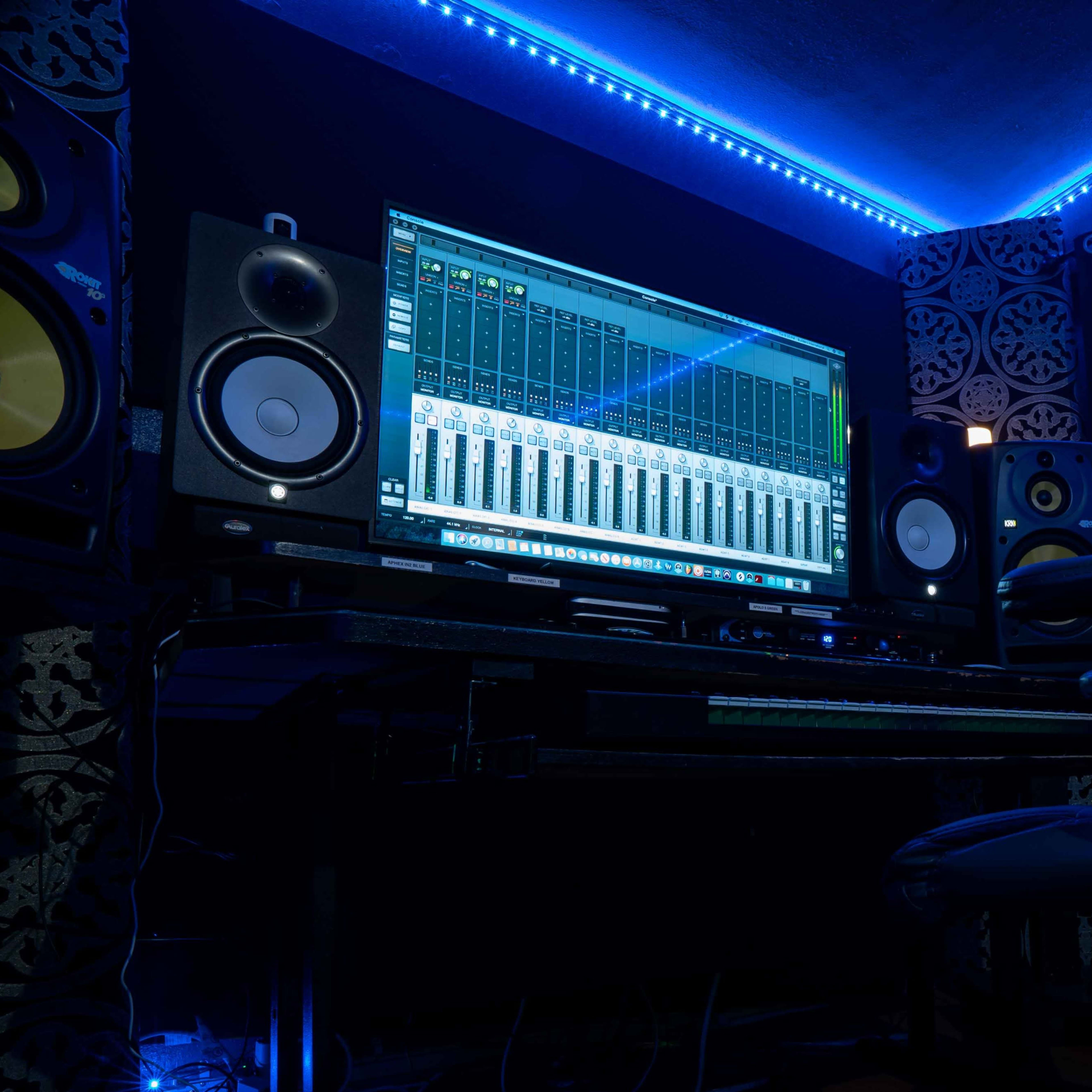 A home recording studio features a computer screen displaying audio mixer software, flanked by studio monitors and illuminated by blue LED lights.