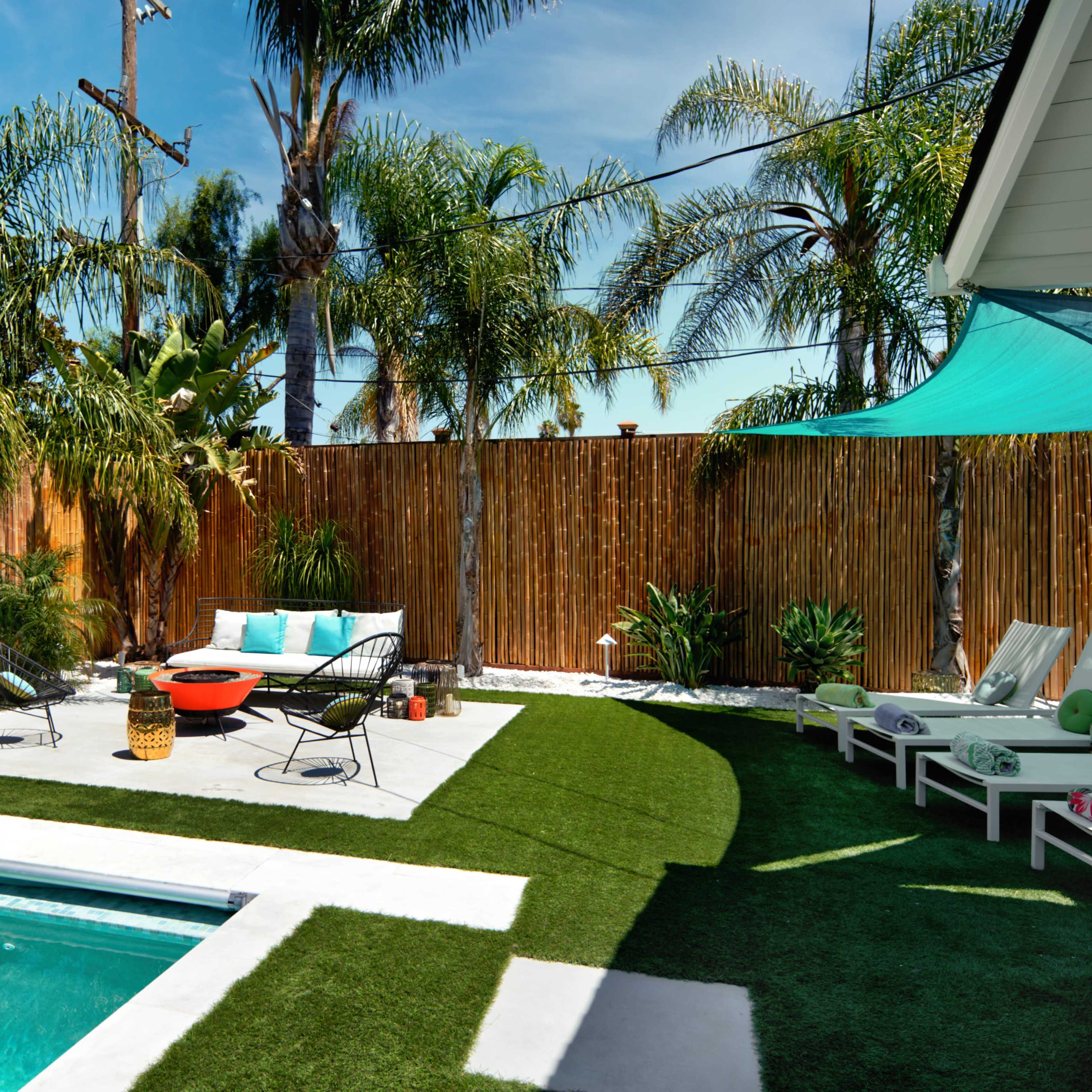 A modern backyard featuring a swimming pool, a seating area with a sofa and chairs, and tropical plants surrounded by a wooden fence.